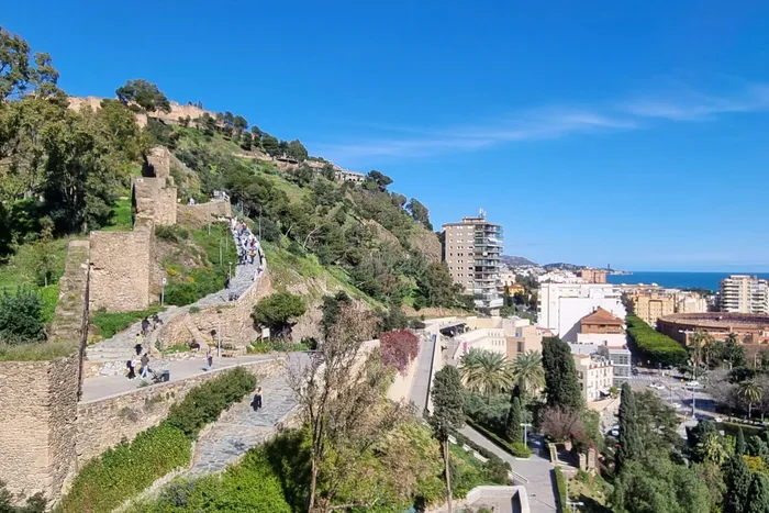 Blick über Málaga mit mittelalterlicher Burgmauer im Vordergrund und moderner Bebauung sowie dem Meer im Hintergrund. Menschen sind auf den Wegen zu sehen.
