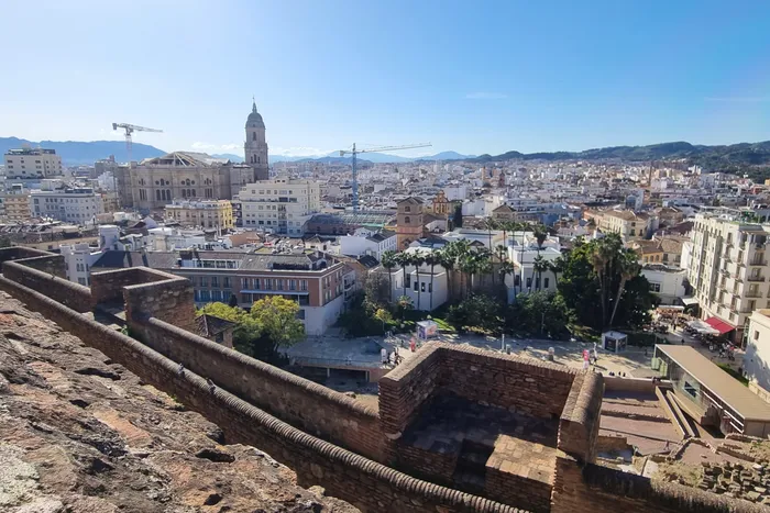 Blick über die Dächer von Málaga mit dem historischen Stadtzentrum und dem Hafen im Hintergrund. Im Vordergrund eine alte Stadtmauer.