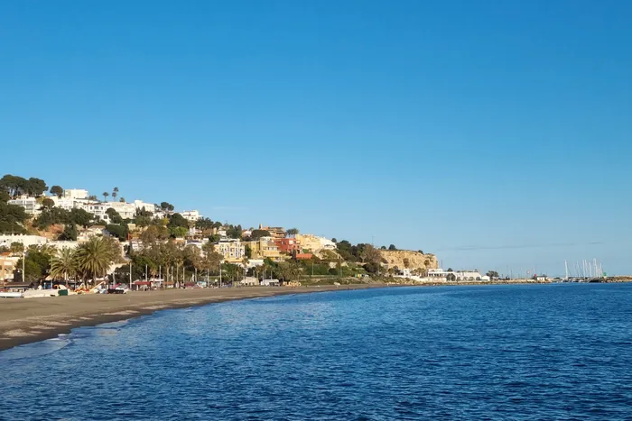 Blick auf die Küstenlinie von Málaga mit Strand, Gebäuden und Palmen unter blauem Himmel. Im Hintergrund sind Jachten im Hafen zu sehen.