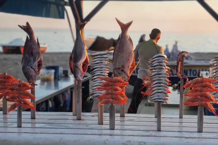 Auf Spieße gesteckte Fische und Garnelen werden in einem Küstenrestaurant präsentiert. Im Hintergrund ist eine Person zu sehen, vermutlich ein Mitarbeiter des Restaurants.