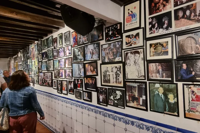 Eine Wand ist mit zahlreichen gerahmten Schwarzweißfotos und Postern bedeckt, darunter Porträts von Personen und Filmszenen. Im Vordergrund steht eine Person mit rotem Oberteil und Jeansjacke, die an der Wand entlanggeht.