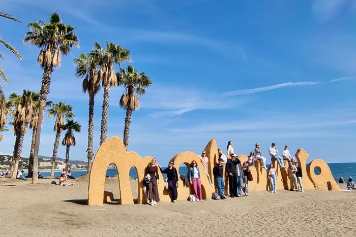Eine Gruppe junger Menschen steht vor einer großen, sandfarbenen Buchstabeninstallation am Strand von Málaga. Im Hintergrund Palmen und das Mittelmeer.