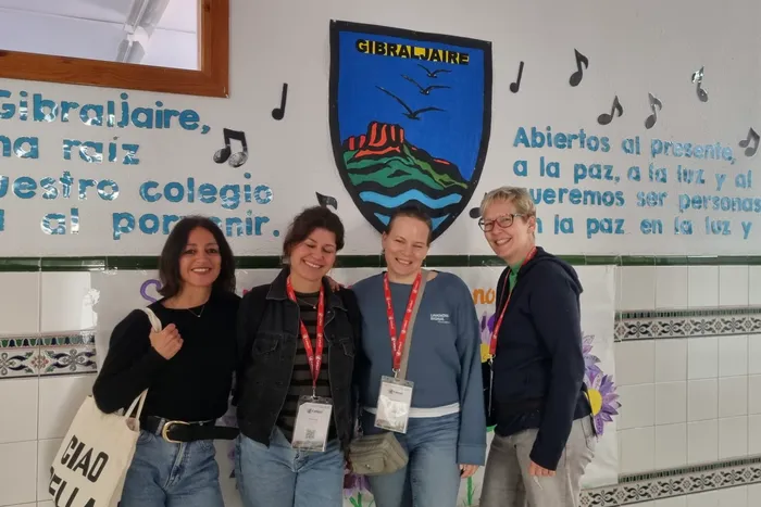 Vier lächelnde Frauen stehen vor einer bemalten Wand mit dem Schriftzug „Gibraltaire“ und musikalischen Noten. Sie tragen Namensschilder und wirken wie Teilnehmerinnen eines Programms in Málaga.