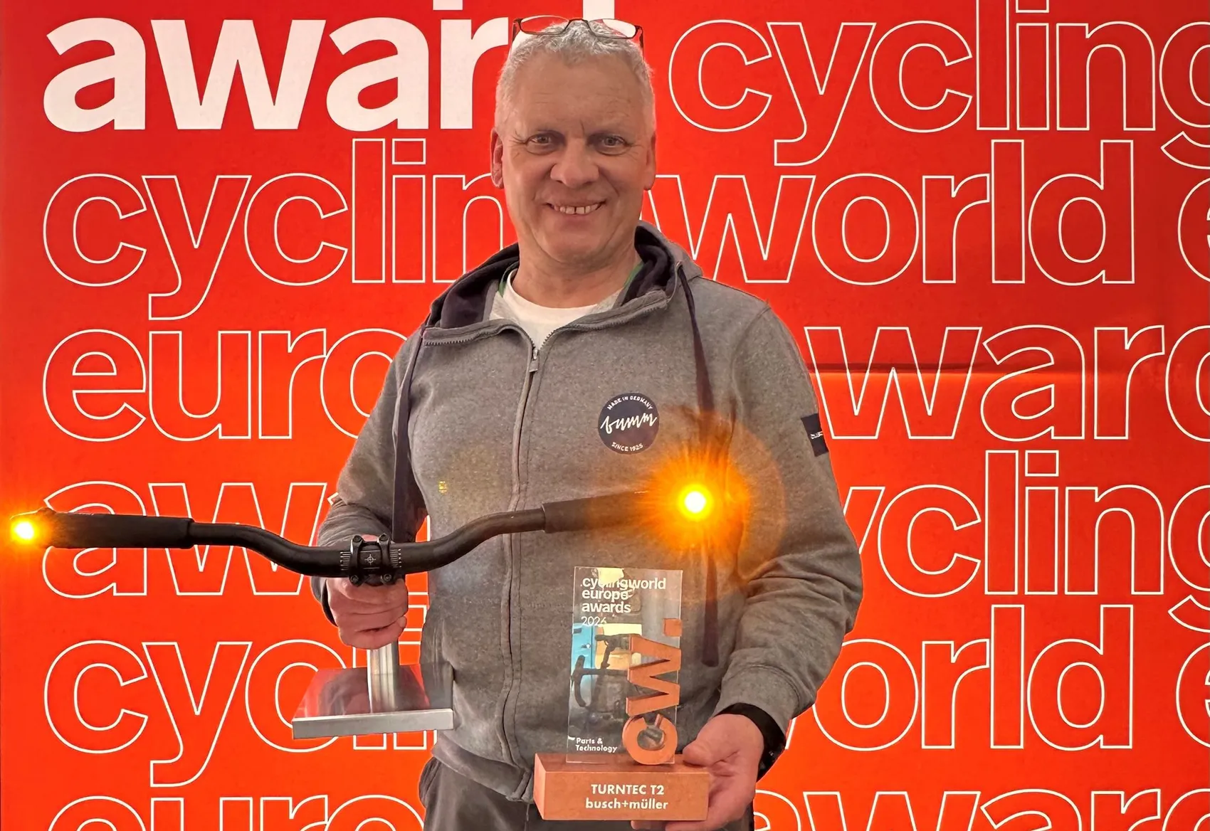 Frank Regge lächelt mit einem Preis in der Hand vor einer Wand mit dem Aufdruck „Cycling World Awards“.