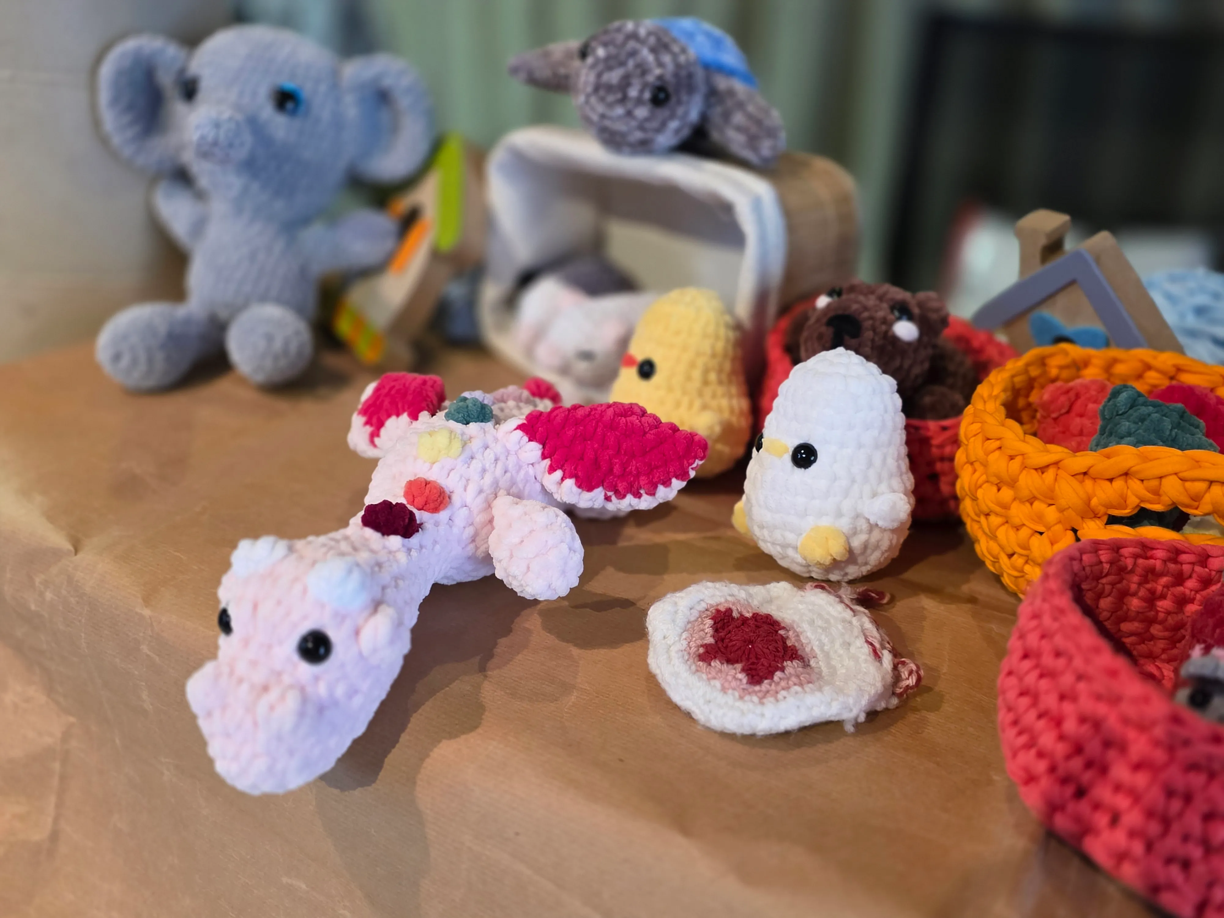 Gehäkelte Stofftiere in Form von Elefant, Nilpferd, Seepferdchen, Küken und weiteren Figuren stehen auf einem Holztisch neben kleinen Körben.