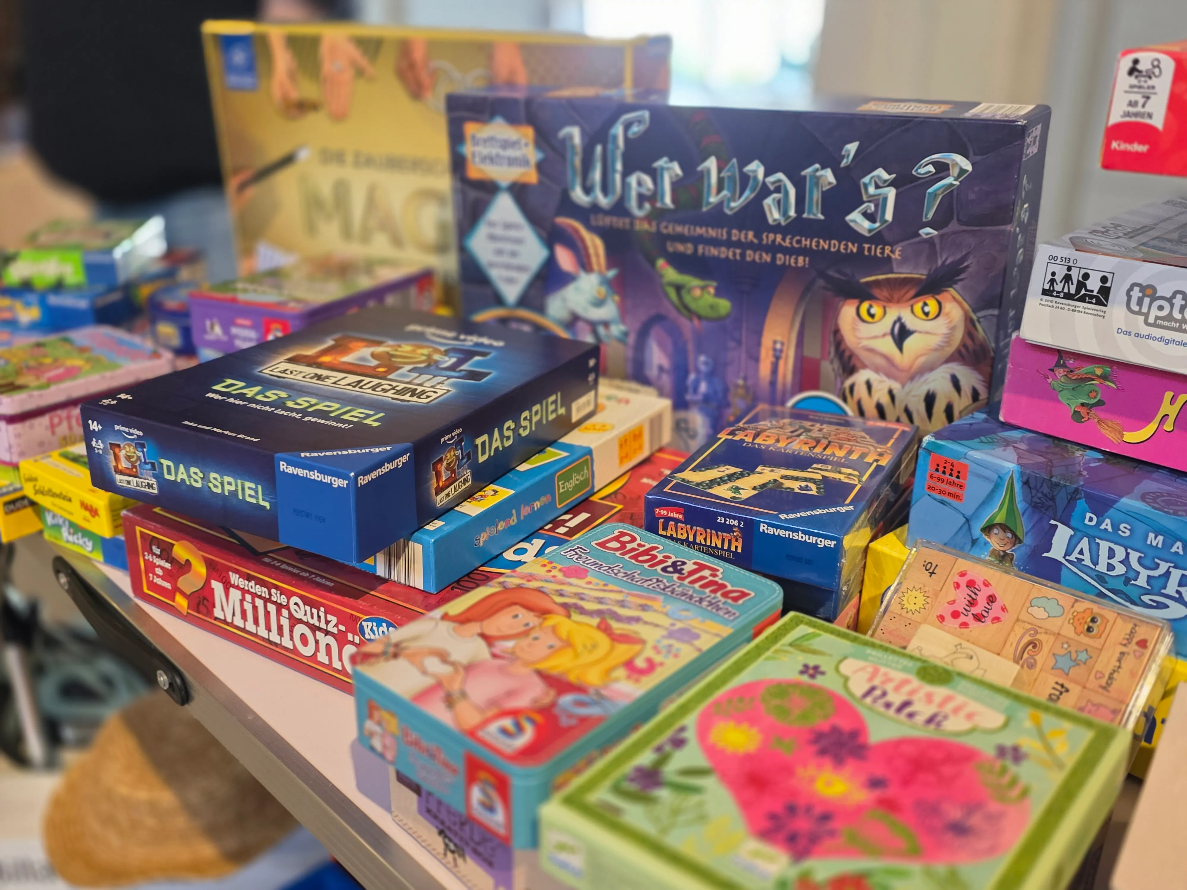 Gestapelte Spielschachteln verschiedener Hersteller und Motive auf einem Holztisch, darunter 'Wer war?' und 'Das Spiel'.