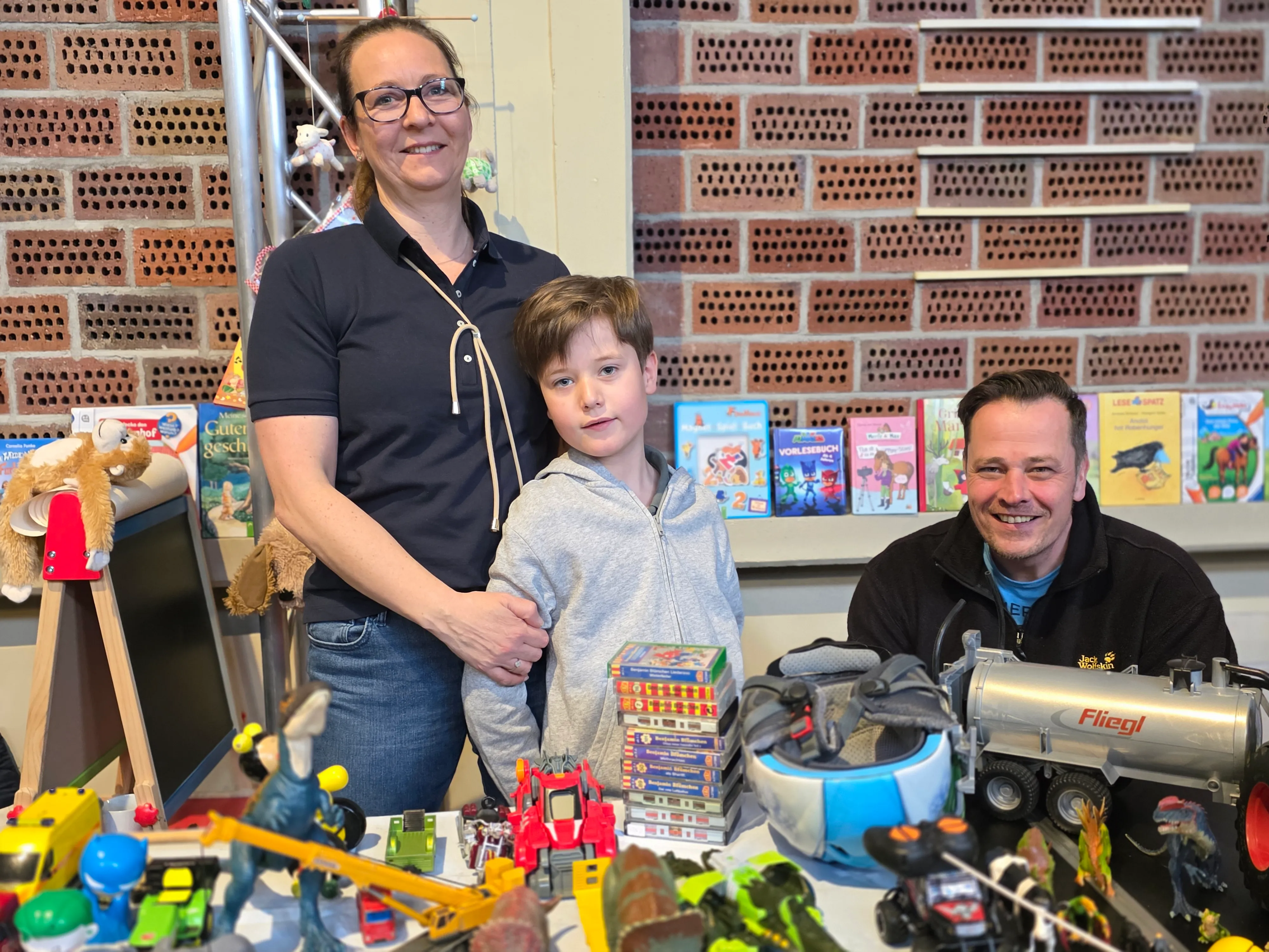 Frau, Junge und Mann stehen vor Regalen mit Büchern und Spielzeug. Im Vordergrund sind viele Spielsachen ausgelegt.