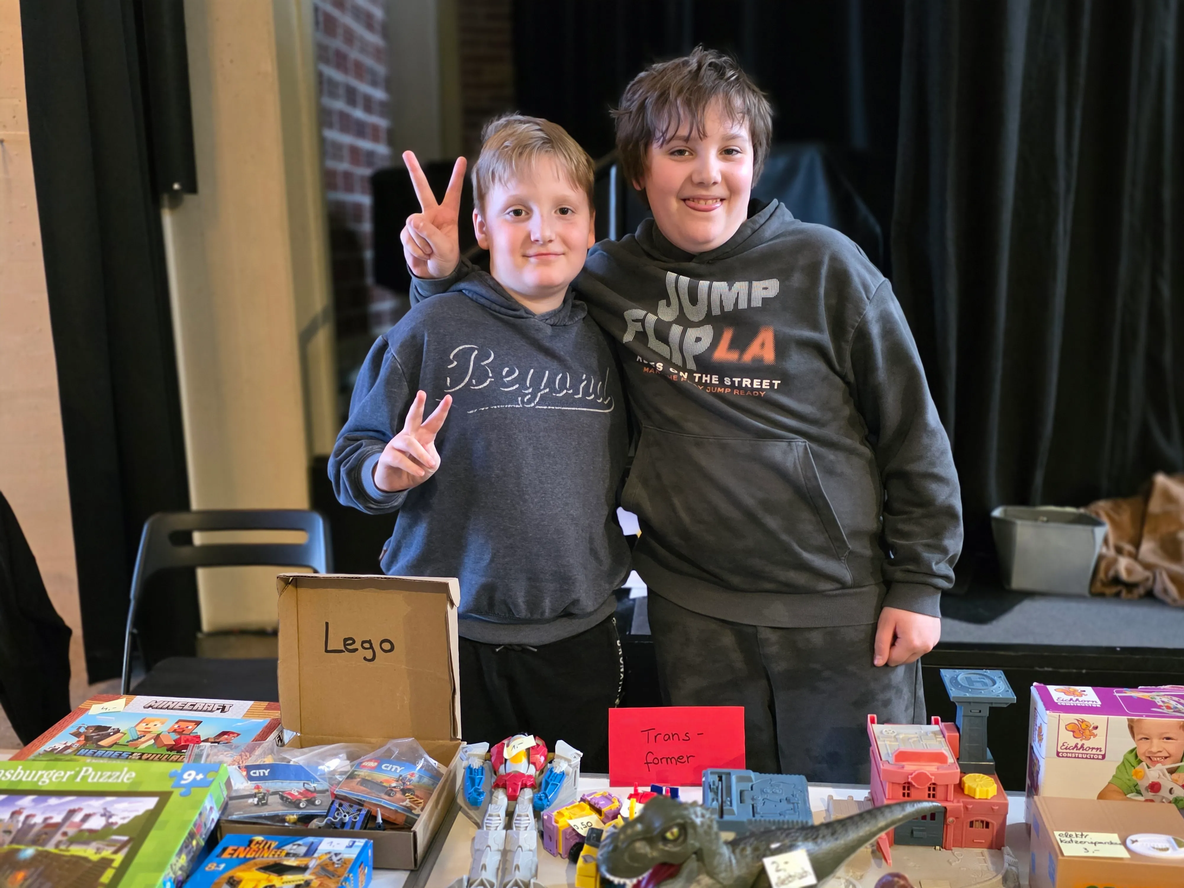 Zwei lächelnde Jungen zeigen den Peace-Zeichen vor einem Tisch voller Spielzeug, darunter Lego und Transformers.