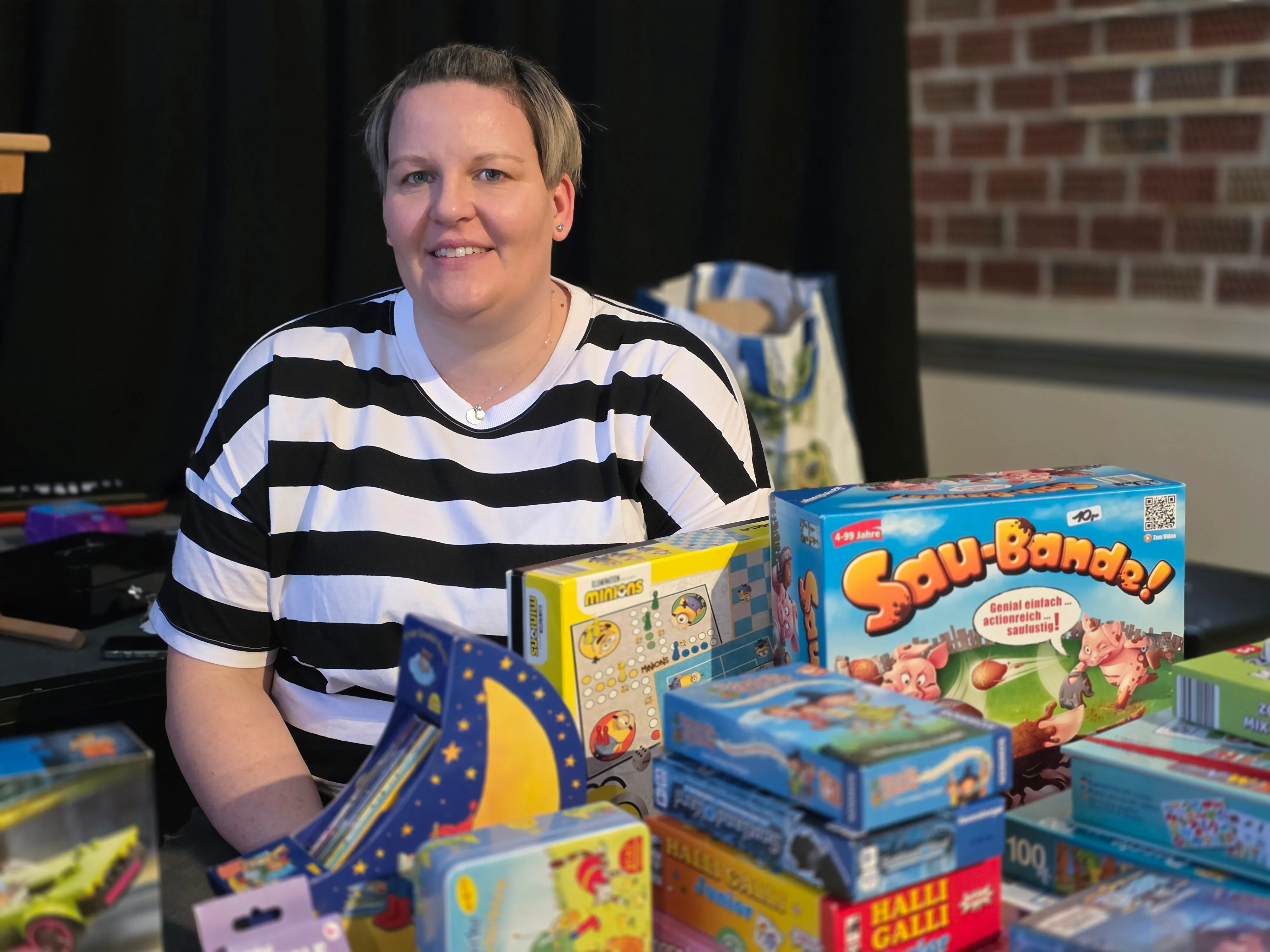 Frau mit kurz geschnittenen, blonden Haaren lächelt vor einem Tisch voller Gesellschaftsspiele, darunter „Minions“ und „Sau-Bande“.