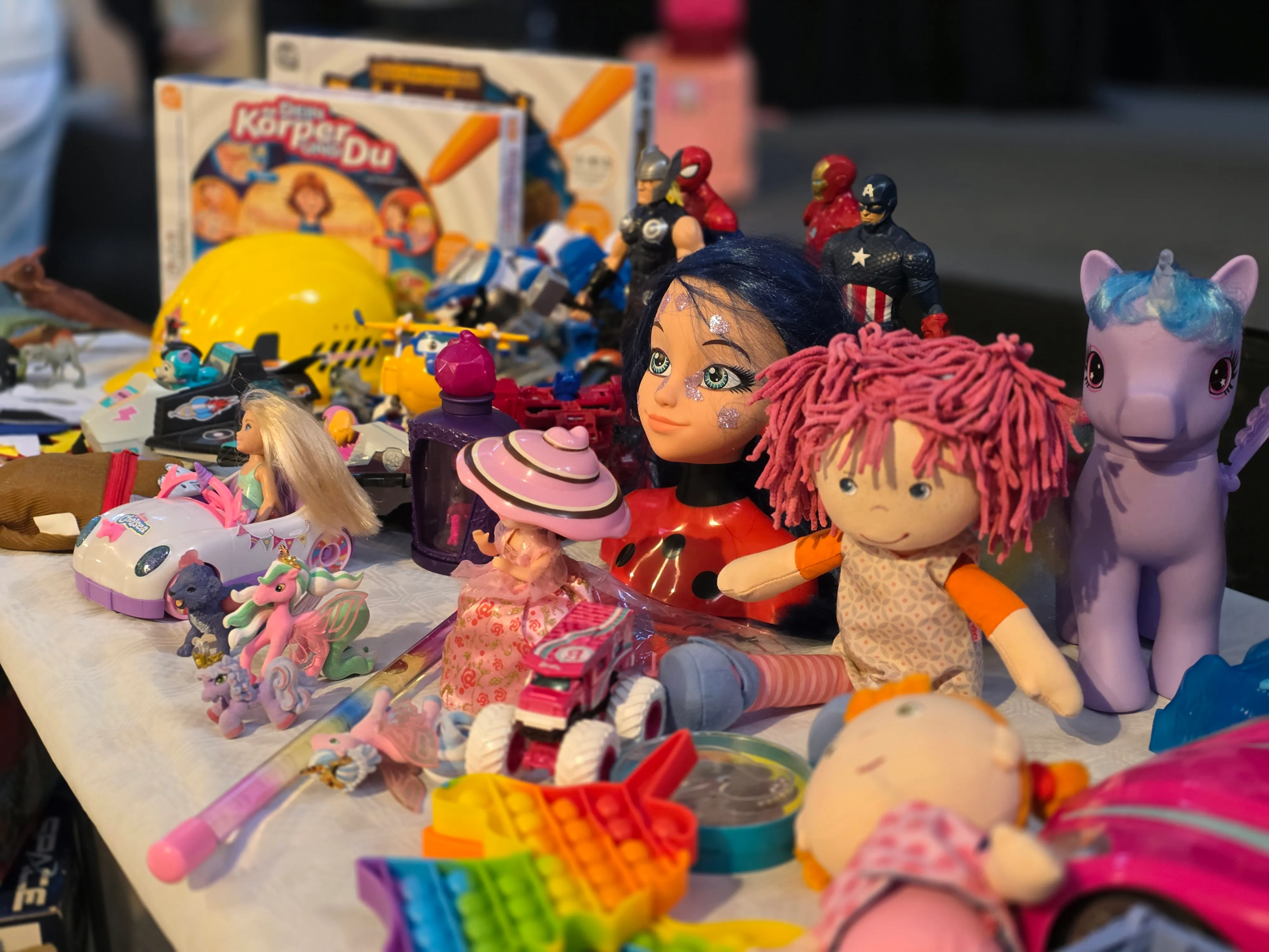 Verschiedenes Spielzeug liegt auf einem Tisch: Puppen, Actionfiguren, ein Spielzeugpferd und Gesellschaftsspiele. Im Hintergrund ist eine Verpackung für das Spiel 'Dein Körper, du' zu sehen.