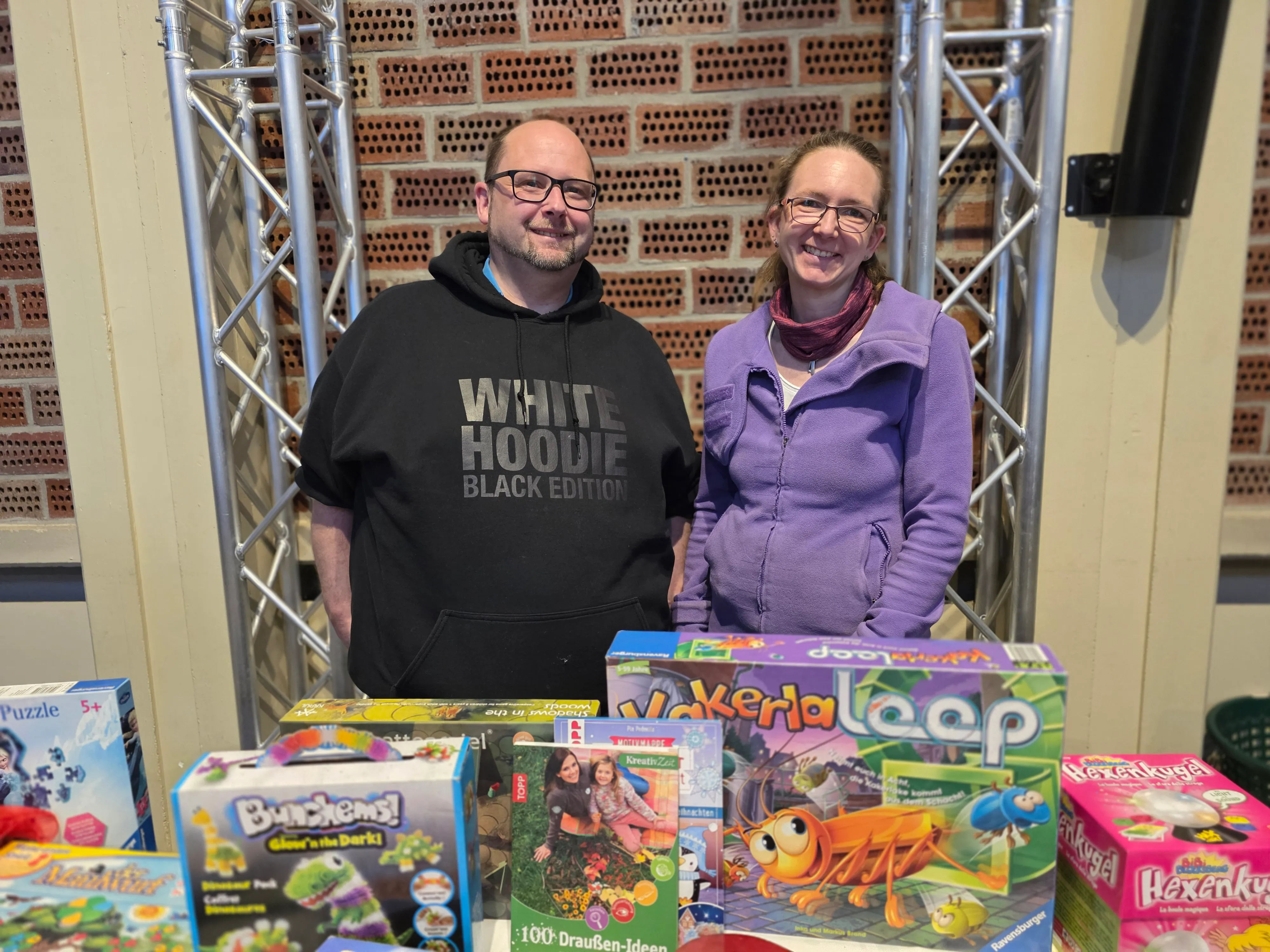 Zwei Personen stehen vor einem Tisch mit Spielzeug. Der Mann trägt ein orangefarbenes T-Shirt mit Aufdruck, die Frau eine lilafarbene Jacke. Im Hintergrund ist ein roter Backsteinbogen zu sehen.