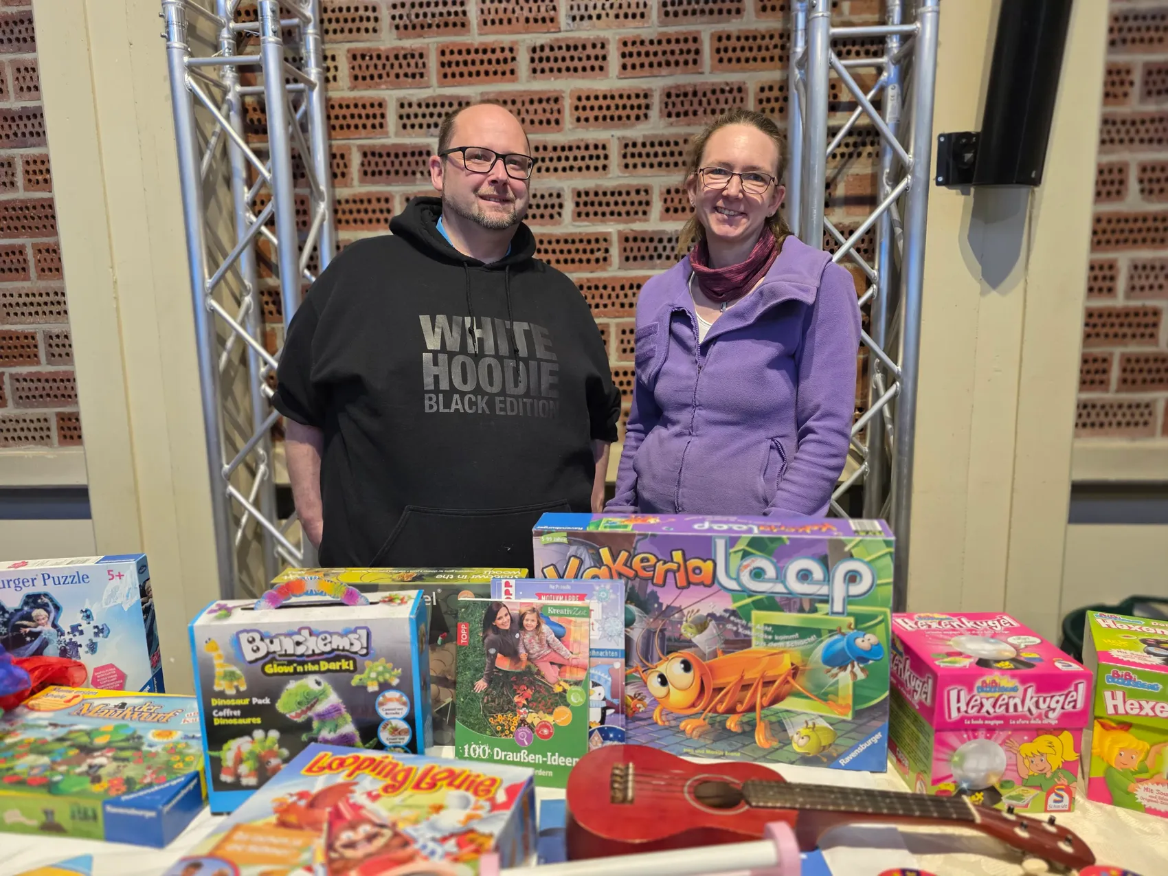 Zwei Personen stehen hinter einem Tisch voller Spielzeug. Im Hintergrund eine Backsteinwand und silberne Metallgerüste.