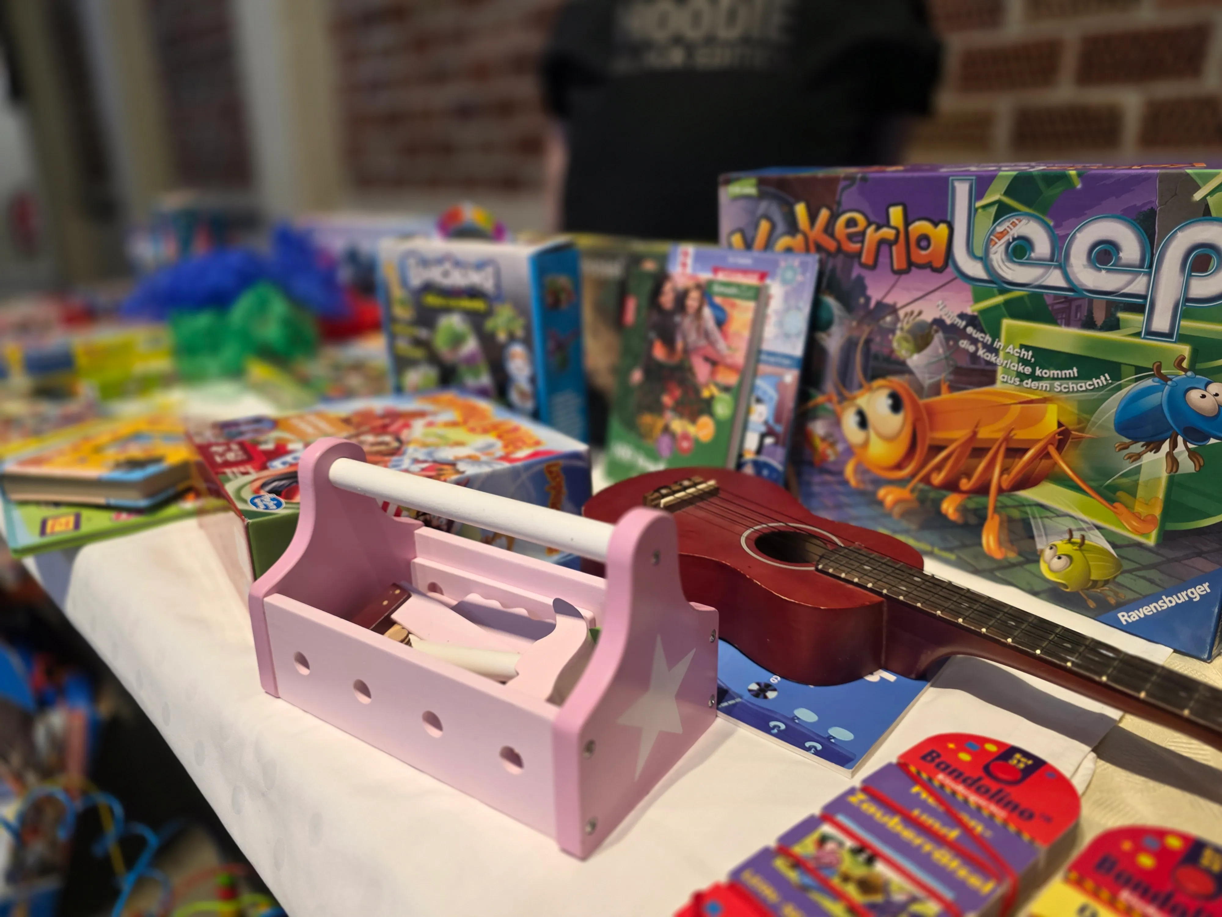 Verschiedene Spielsachen und Spiele stehen auf einem Tisch, darunter ein pinkfarbenes Kinderstuhl und das Spiel 'Kalerialeep' mit einem bunten Dinosaurier auf der Verpackung.