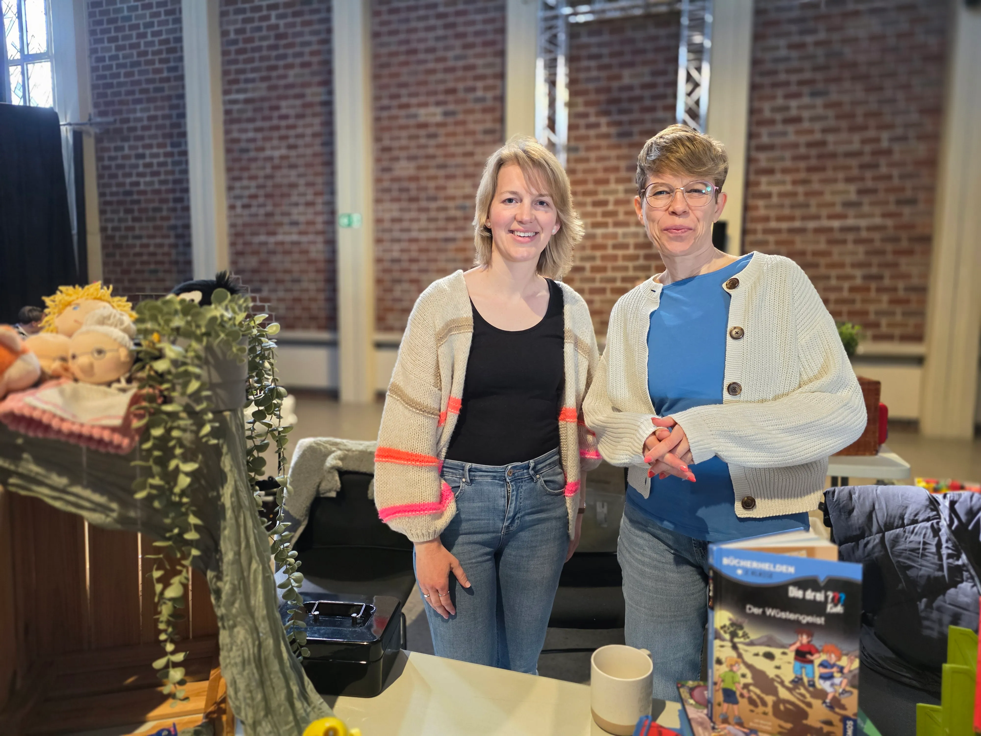 Zwei lächelnde Frauen stehen nebeneinander an einem Tisch mit Büchern und Spielzeug. Im Hintergrund sind Backsteinwände und weitere Tische mit Angeboten zu sehen.