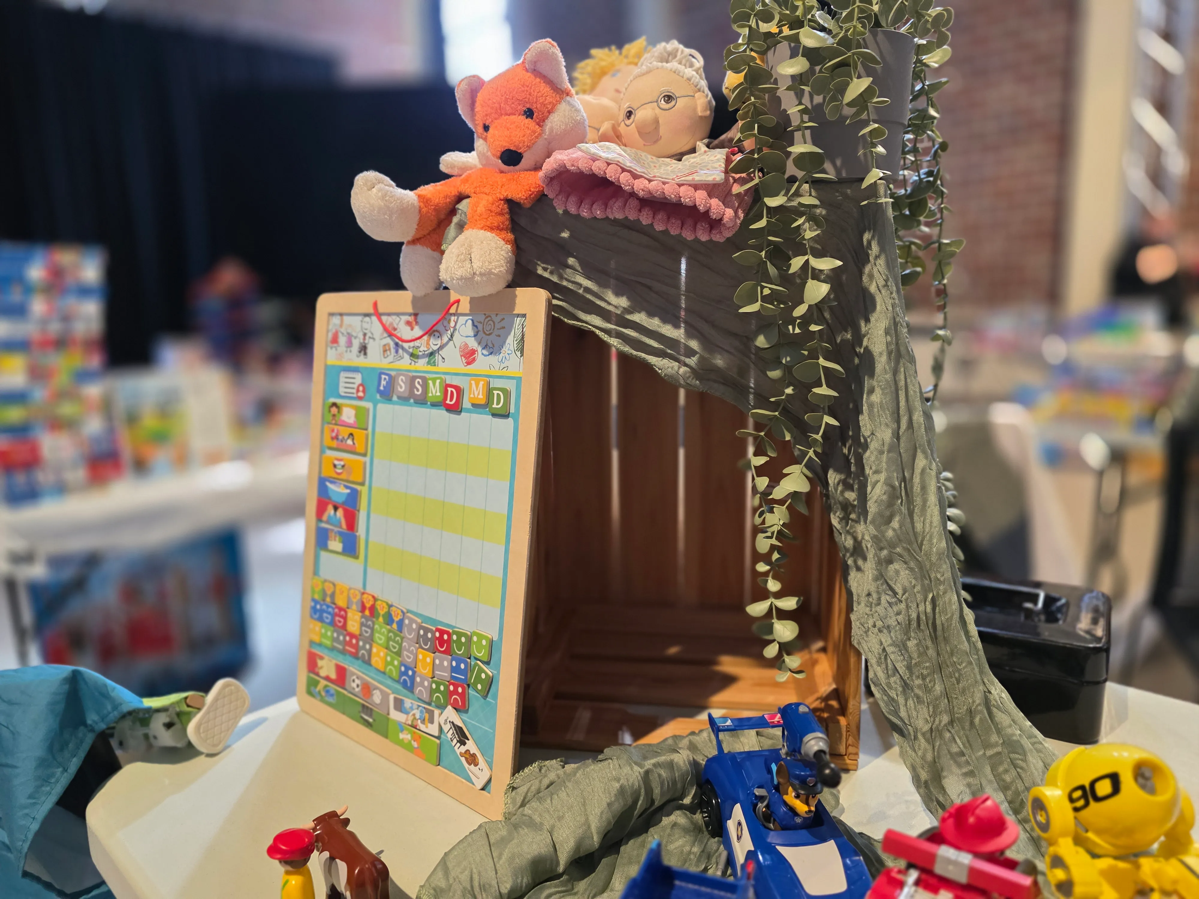 Ein Spielzeughaus mit Stofftieren auf dem Dach und Spielzeugautos davor steht auf einem Tisch. Im Hintergrund sind weitere Spielsachen und Menschen zu sehen.