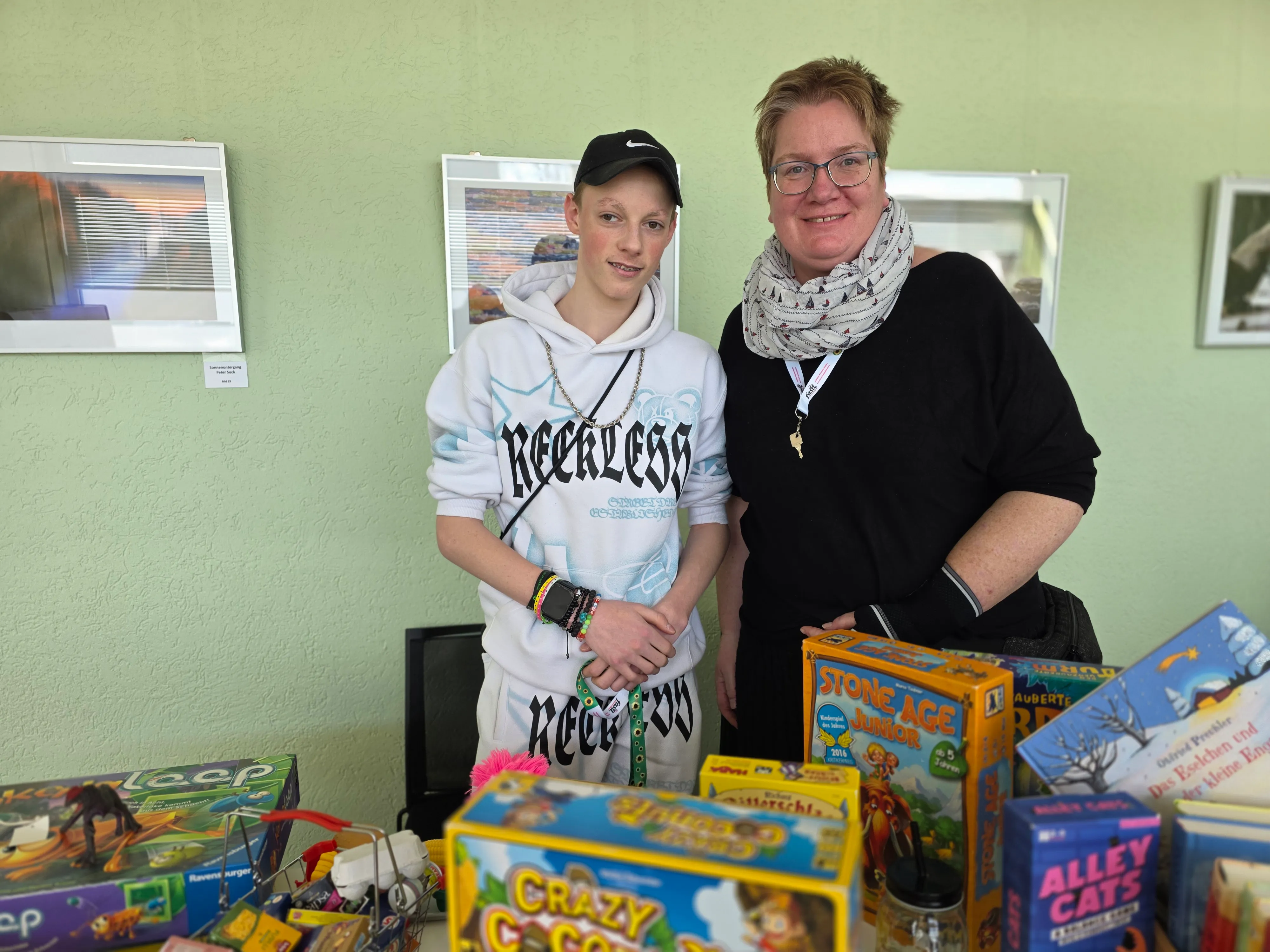 Ein Jugendlicher mit Baseballkappe und eine Frau mit Brille stehen vor einem Tisch voller Gesellschaftsspiele. Im Hintergrund hängt ein gerahmtes Bild an einer hellgrünen Wand.