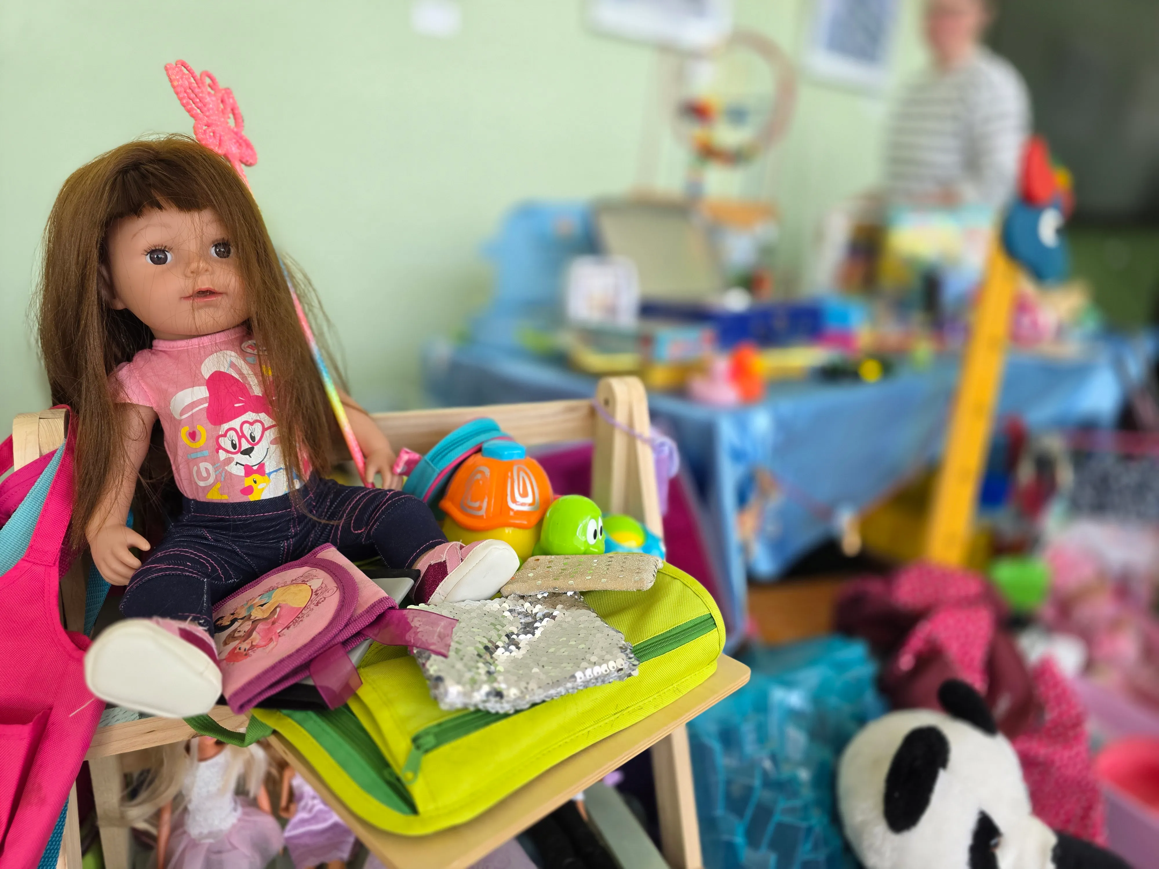 Eine Puppe mit langen, braunen Haaren sitzt auf einem Tisch voller Spielzeug, darunter ein Plüschtier und eine glitzernde Tasche. Im Hintergrund sind weitere Spielsachen und Tische zu sehen.