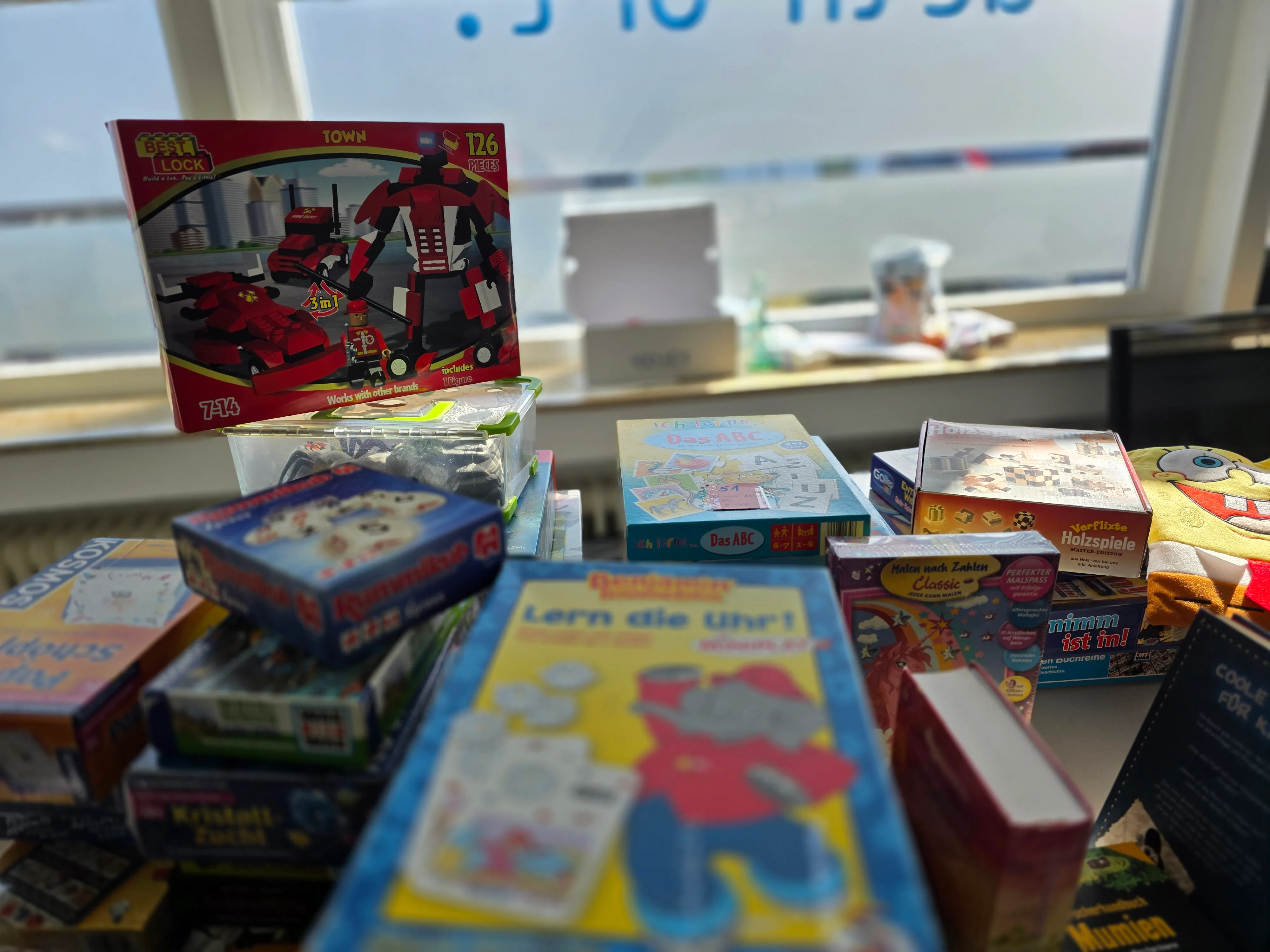 Gestapelte Spielzeugschachteln und ein Roboter-Baukasten stehen auf einem Tisch vor einem Fenster. Im Hintergrund ist eine Person zu sehen.