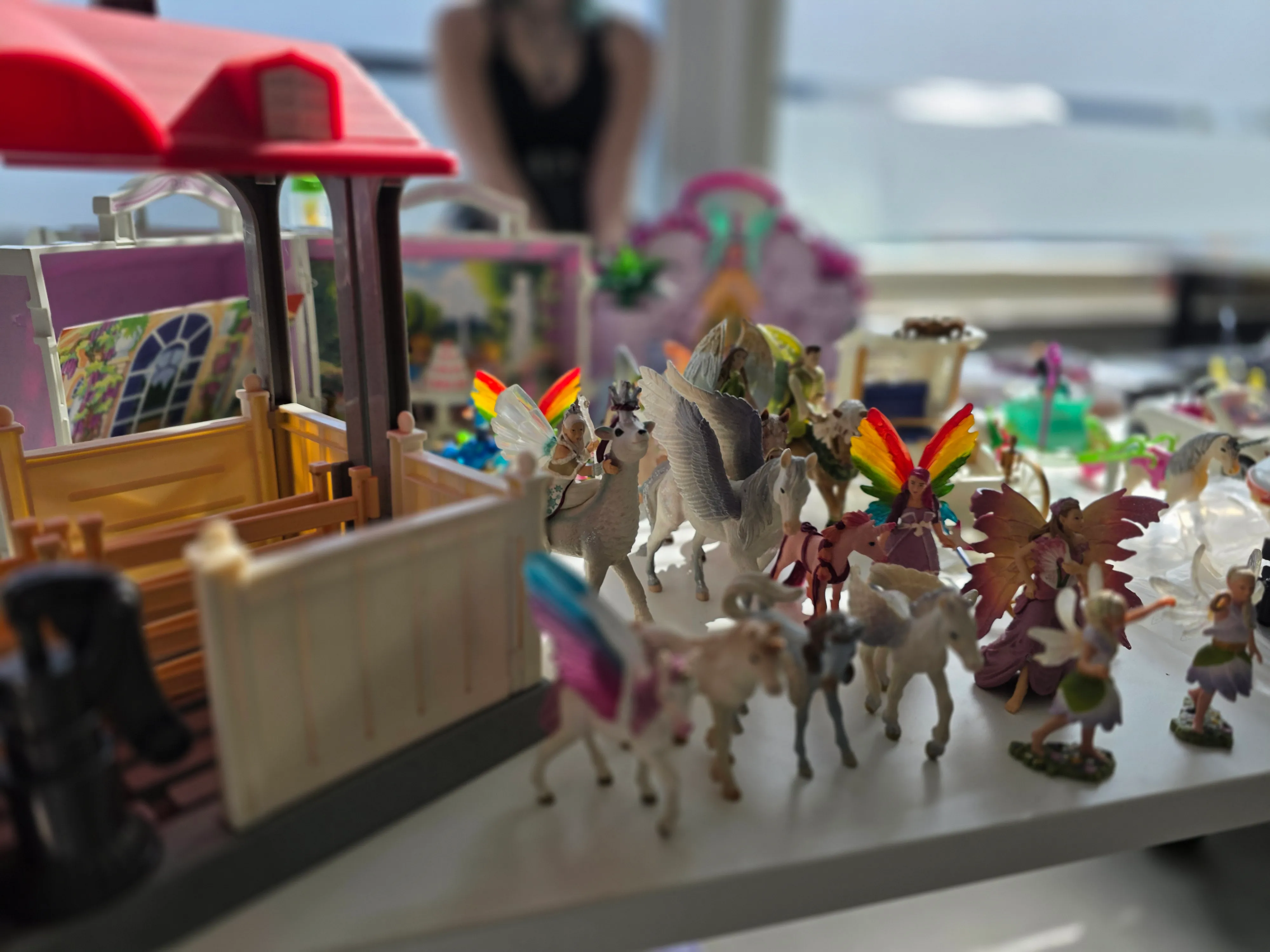 Spielzeugpferde mit geflügelten Figuren und eine Spielzeugfarm stehen auf einem Tisch, im Hintergrund verschwommen weitere Spielsachen und eine Person.