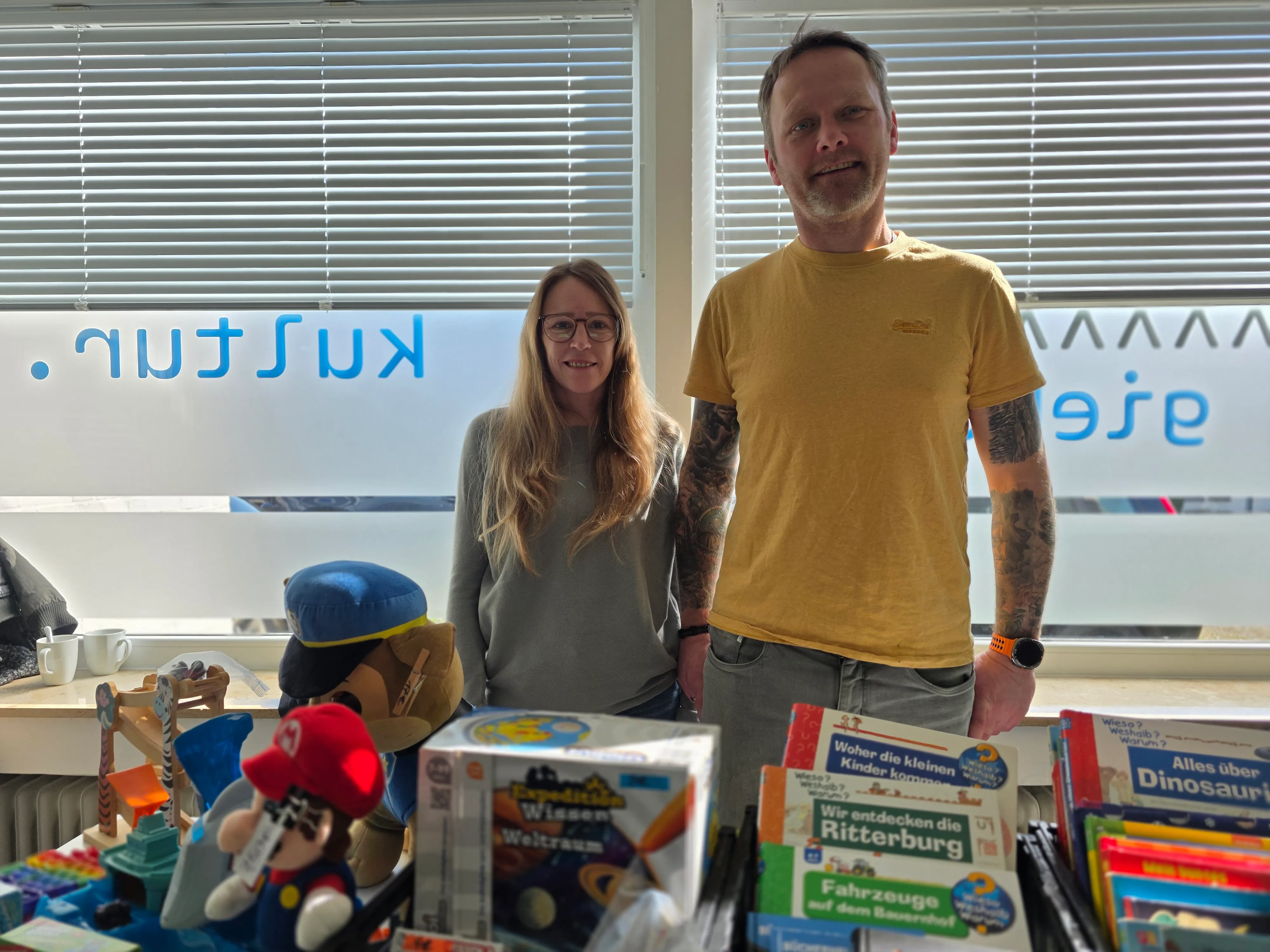 Mann und Frau stehen hinter einem Tisch mit Spielzeug, Büchern und Stofftieren in einem hellen Raum.