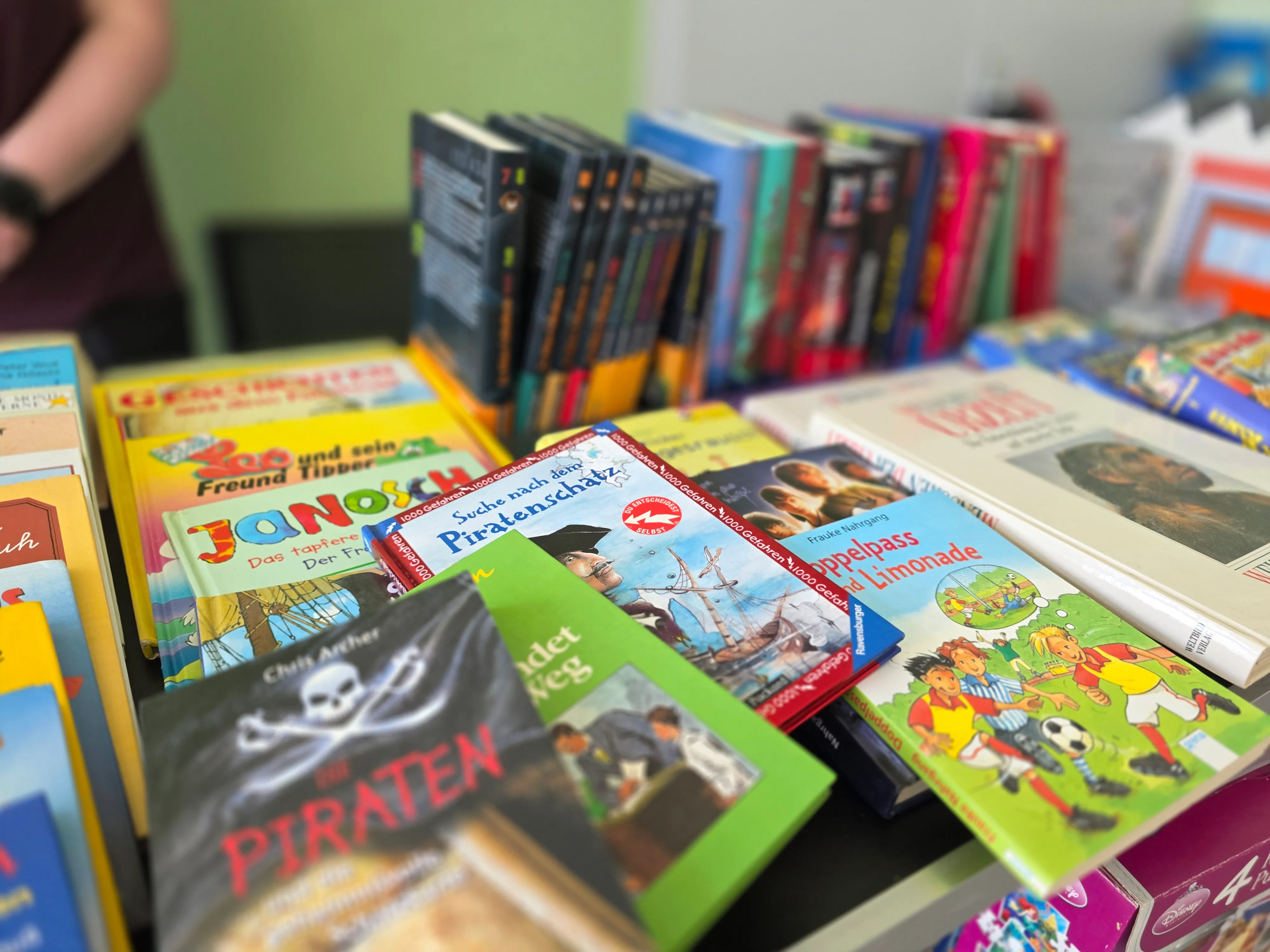 Gestapelte Kinderbücher auf einem Tisch, darunter Titel wie „Janosch“, „Piraten“ und „Doppelpass Limonade“.