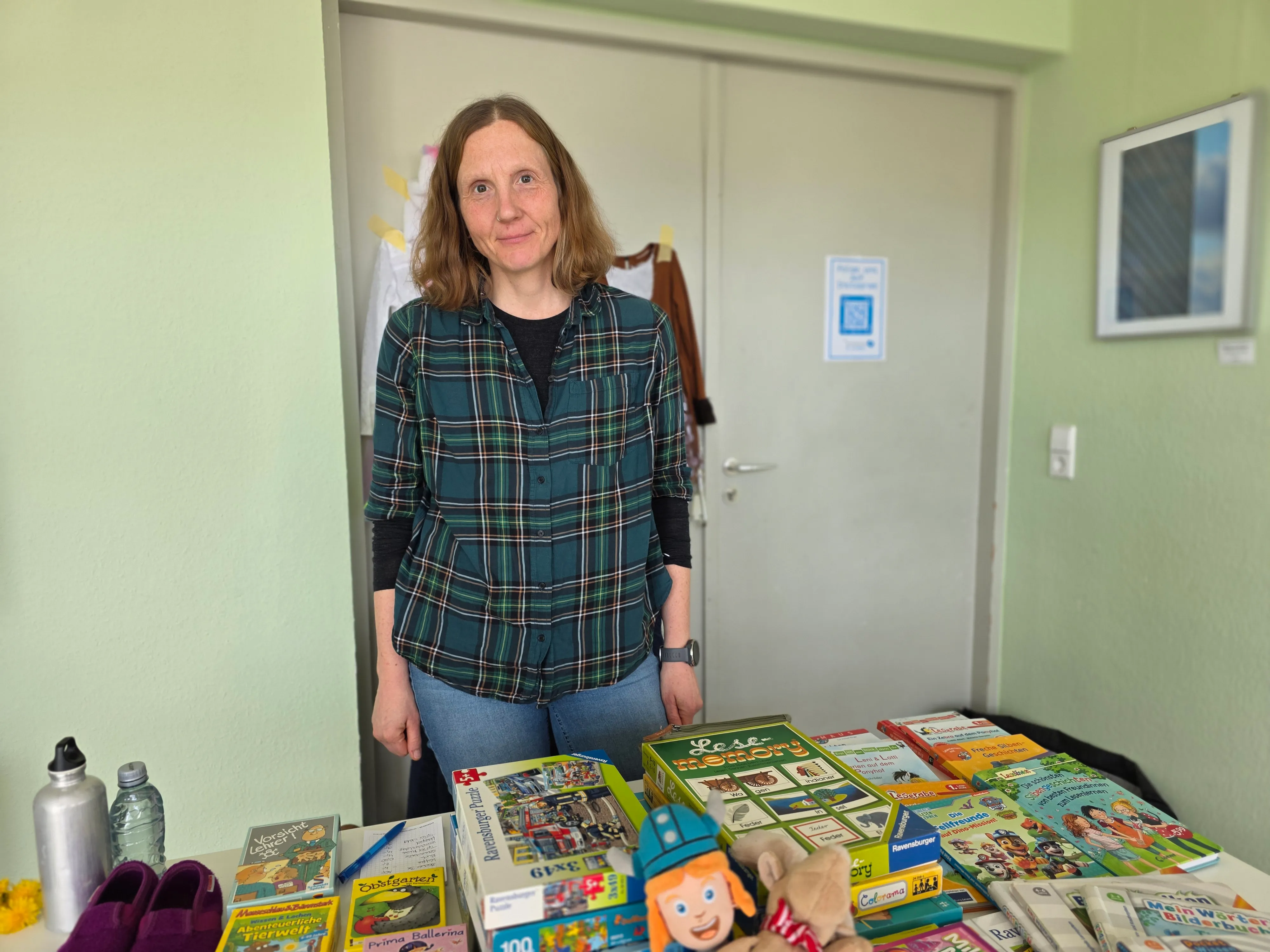 Frau steht lächelnd hinter einem Tisch voller Kinderbücher und Spielzeug in einem hellen Raum.
