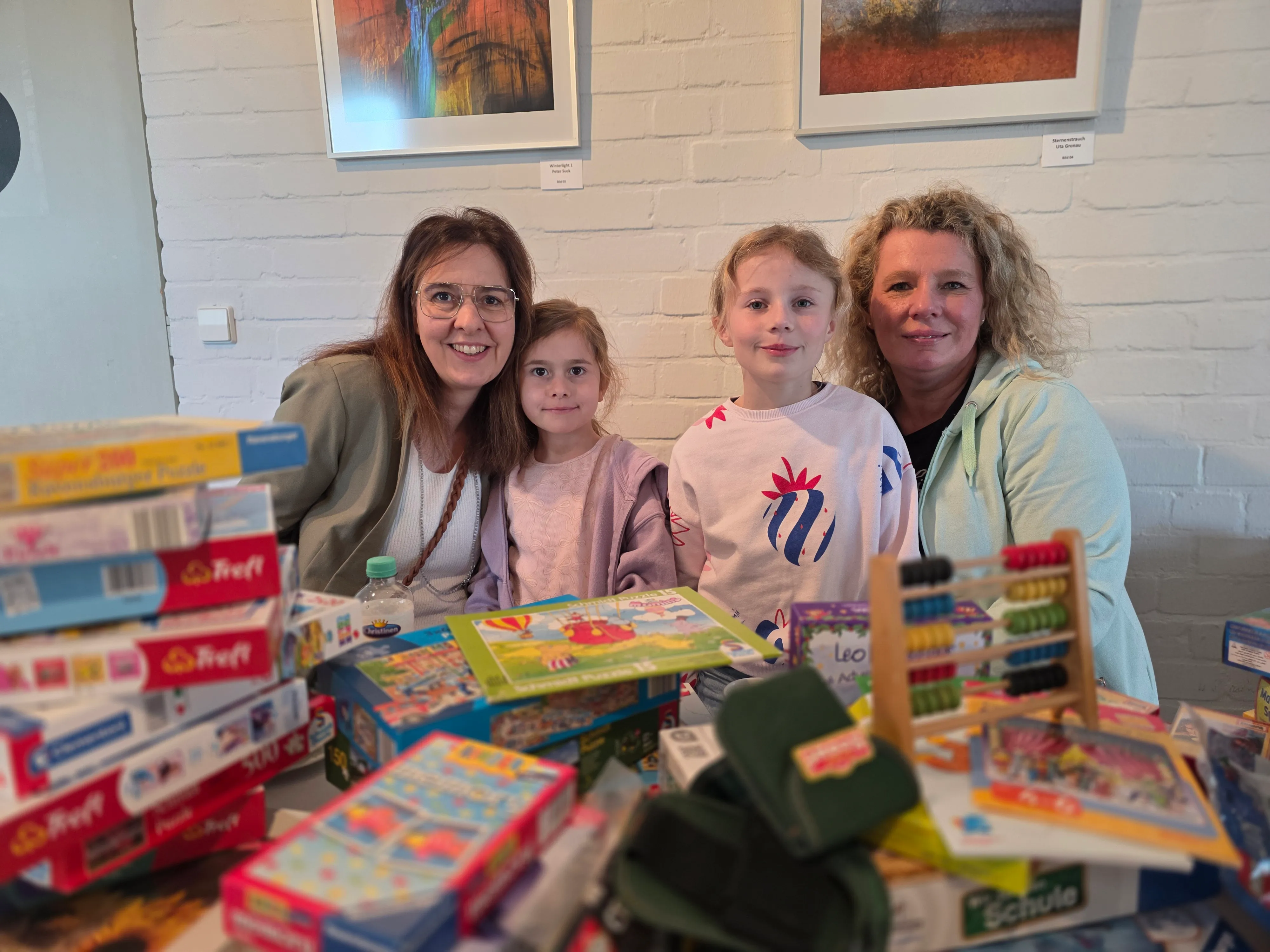 Zwei Frauen und zwei Mädchen stehen vor einem Tisch voller Spielzeug. Im Hintergrund hängen Bilder an einer weißen Backsteinwand.