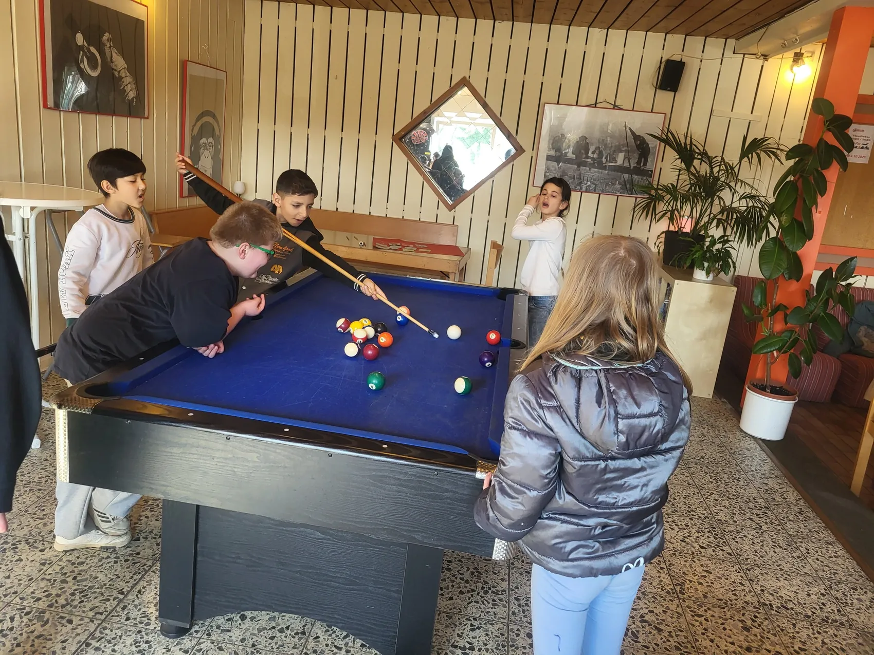 Ein Junge stößt bei einer Billardpartie eine Kugel, während andere Kinder zuschauen. Im Hintergrund hängen Bilder an der Holzwand des Jugendzentrums.