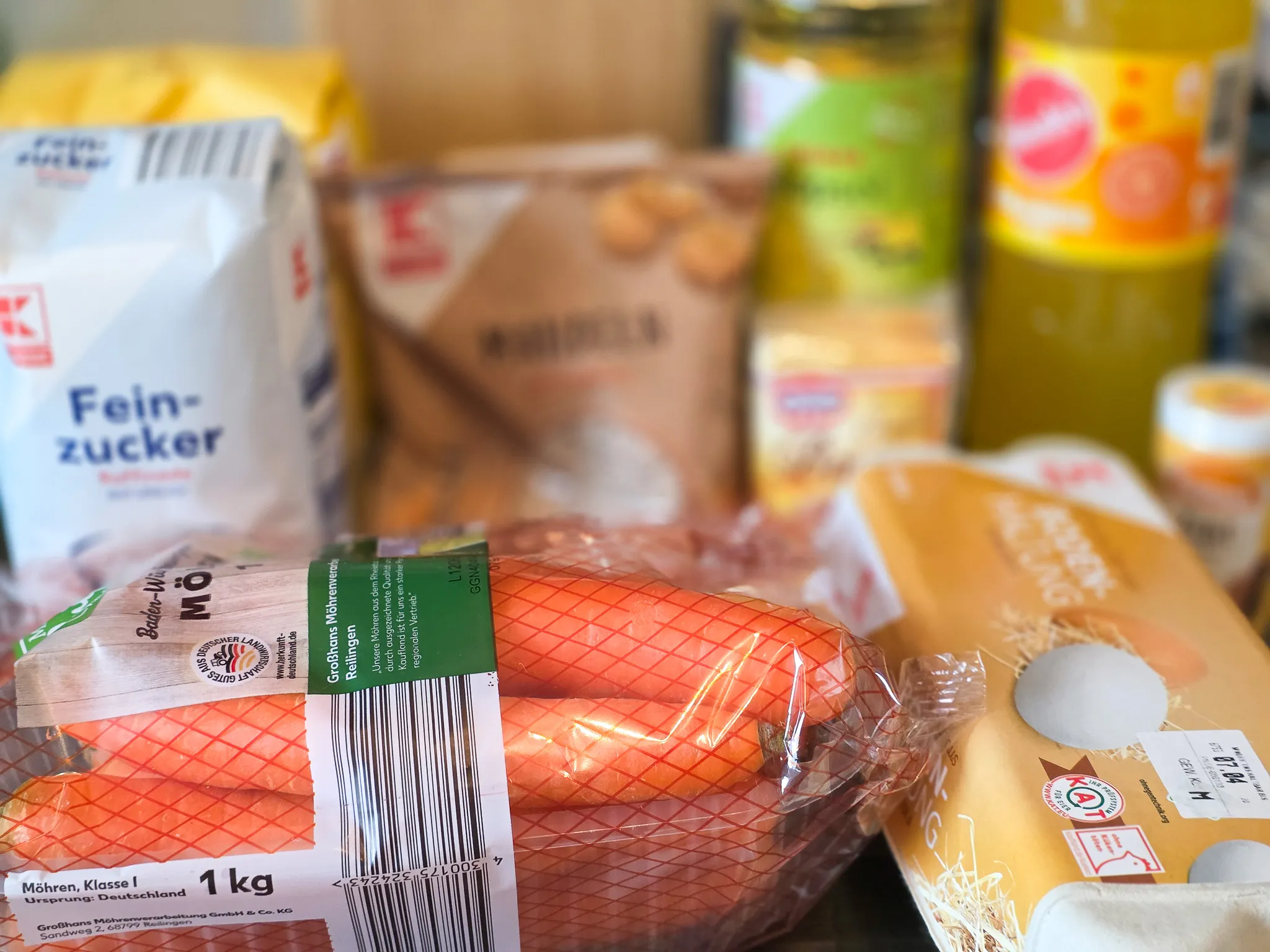 Zucker, Möhren und Reibekuchen in Verpackungen stehen auf einem Holztisch. Im Vordergrund eine Packung Möhren mit dem Hinweis „1 kg“ und „Deutschland“ als Herkunftsland.