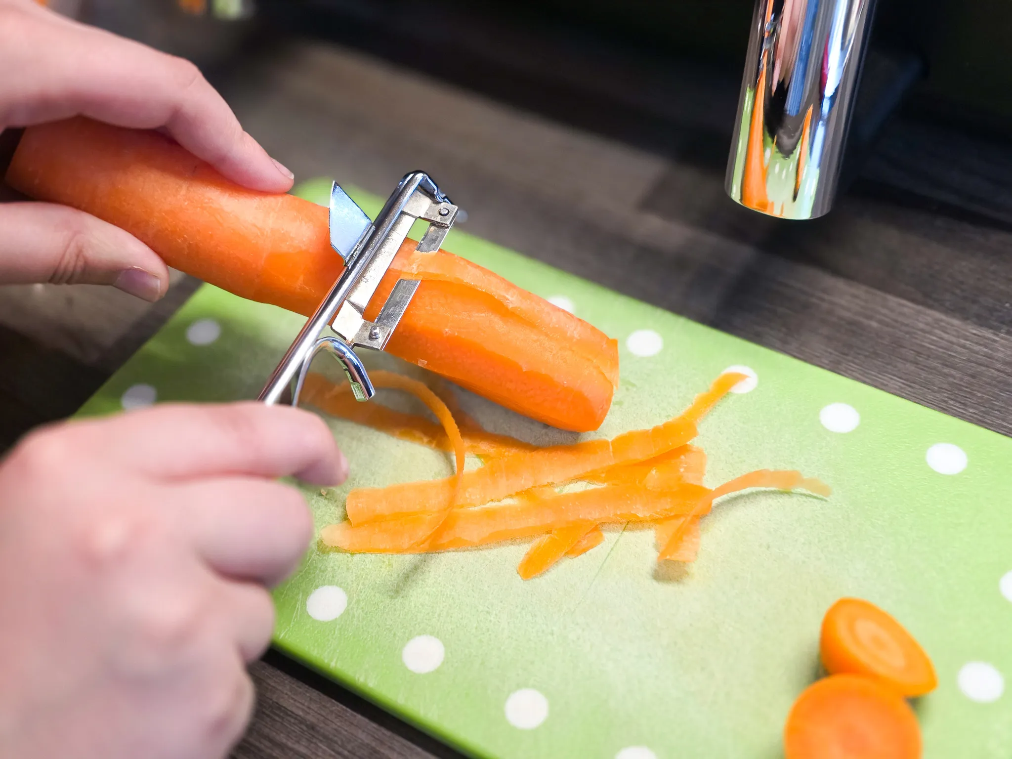 Eine Person schält eine Karotte mit einem Gemüsehobel auf einem grün-weiß gepunkteten Schneidebrett. Geschälte Karottenstreifen und geschnittene Karottenstücke liegen daneben.