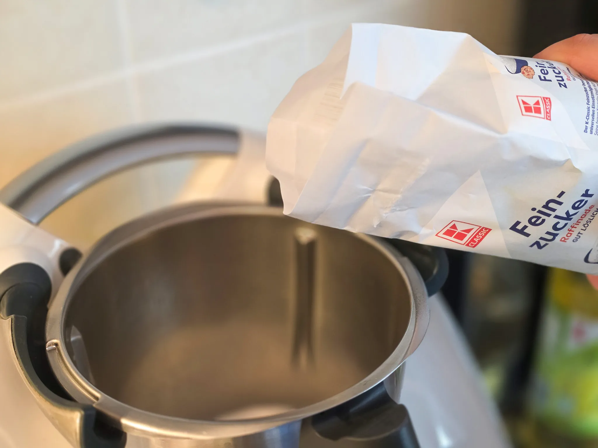 Zucker wird aus einem weißen Beutel in einen großen, silberfarbenen Topf geschüttet.