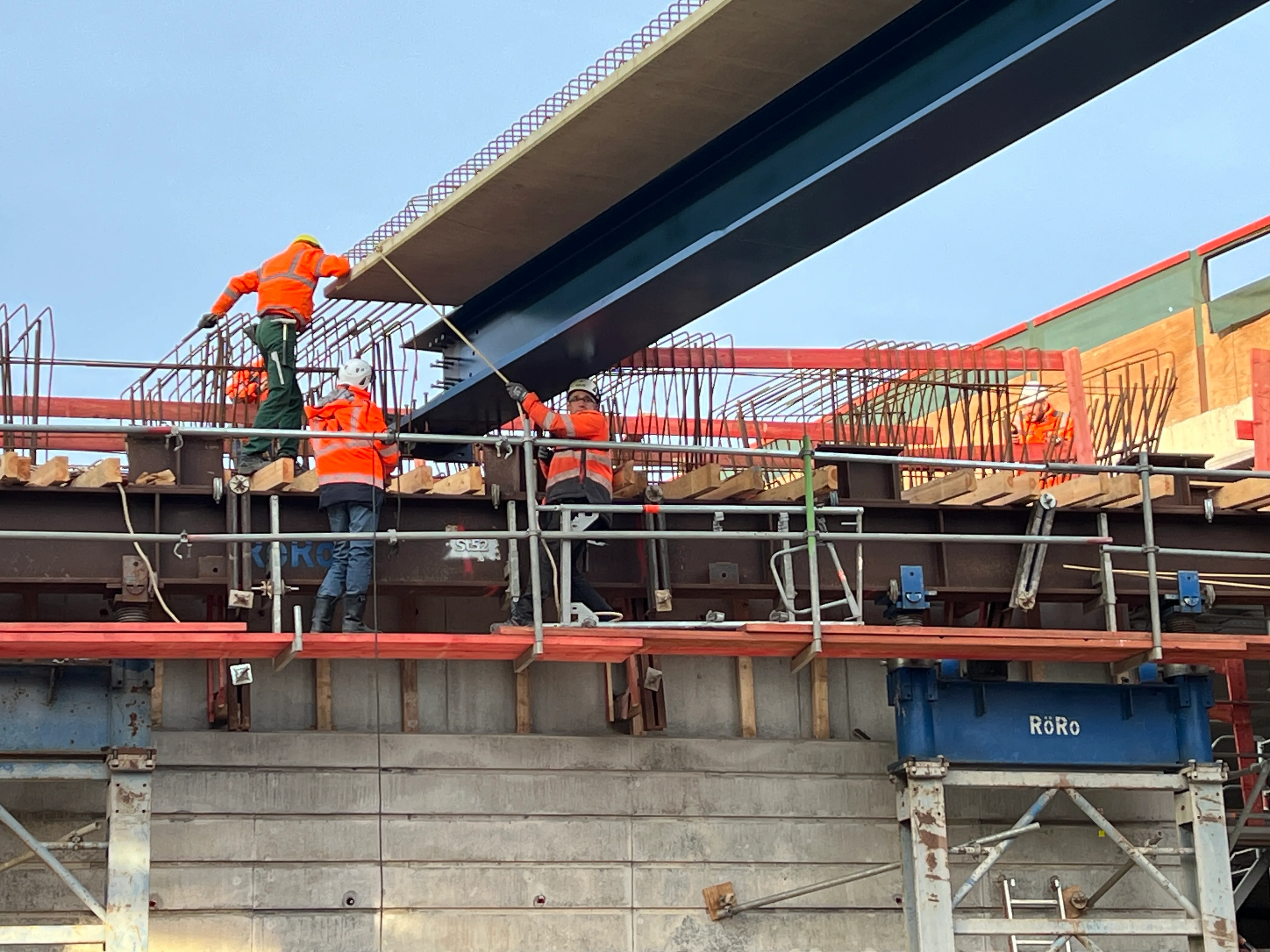 Bauarbeiter in orangefarbenen Westen heben einen großen Stahlträger an, während weitere Arbeiter im Hintergrund auf einem Gerüst arbeiten.