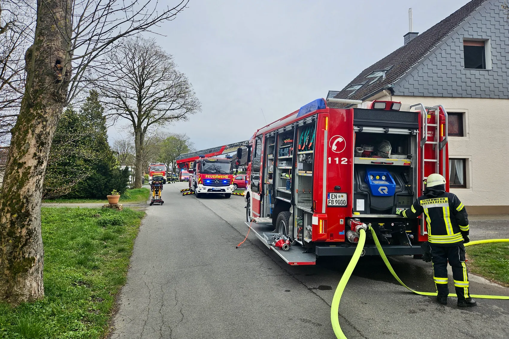 Mehrere Einsatzfahrzeuge der Feuerwehr Breckerfeld stehen vor einem Wohnhaus, während ein Feuerwehrmann einen Schlauch ausrollt.
