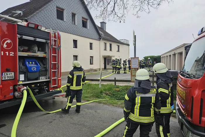 Feuerwehrleute sind bei einem Kaminbrand in Königsheide im Einsatz. Mehrere Einsatzkräfte sind vor einem zweigeschossigen Gebäude zu sehen, aus dem gelbe Schläuche führen.