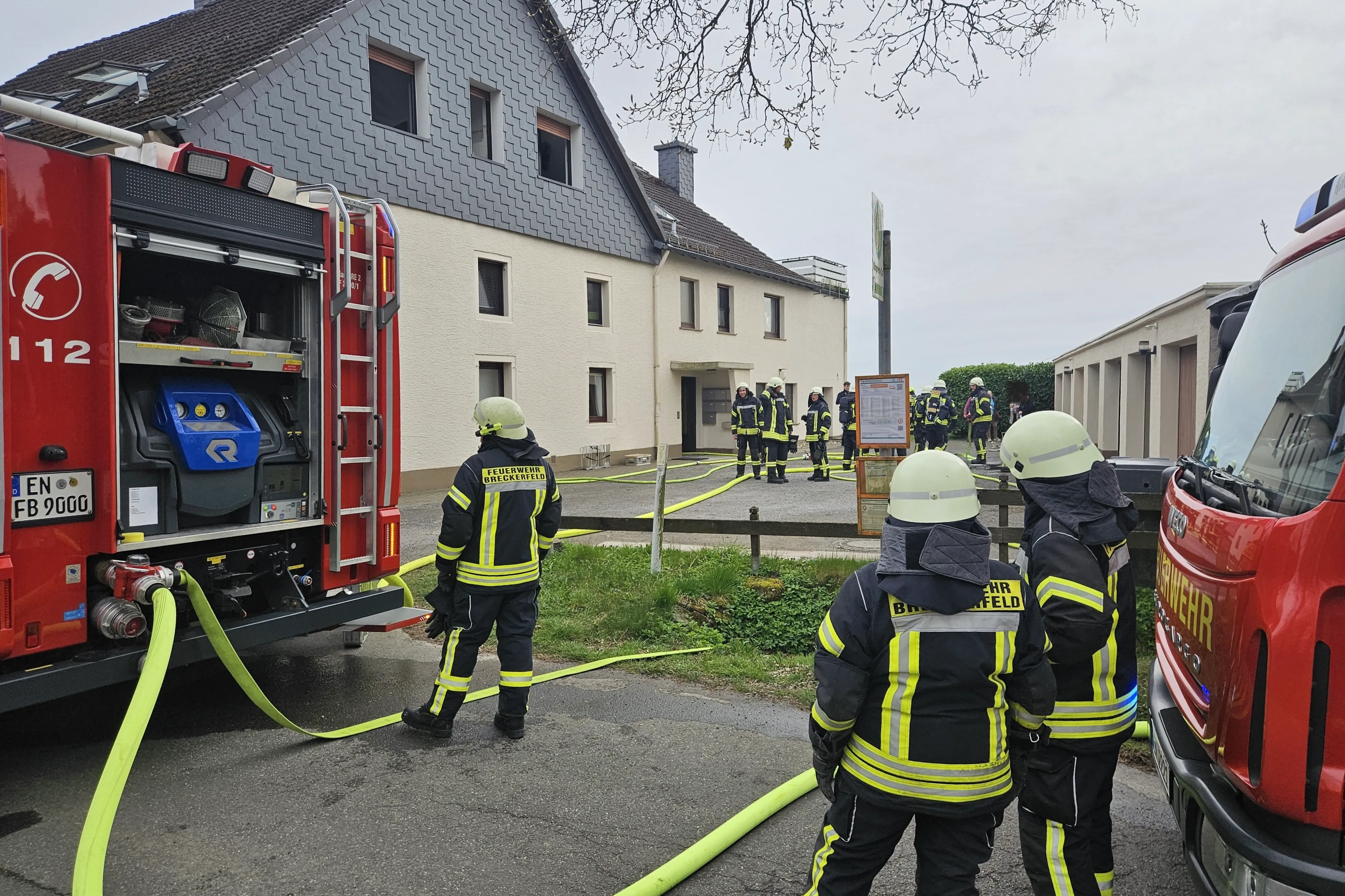 Feuerwehrleute sind bei einem Kaminbrand in Königsheide im Einsatz. Mehrere Einsatzkräfte sind vor einem zweigeschossigen Gebäude zu sehen, aus dem gelbe Schläuche führen.