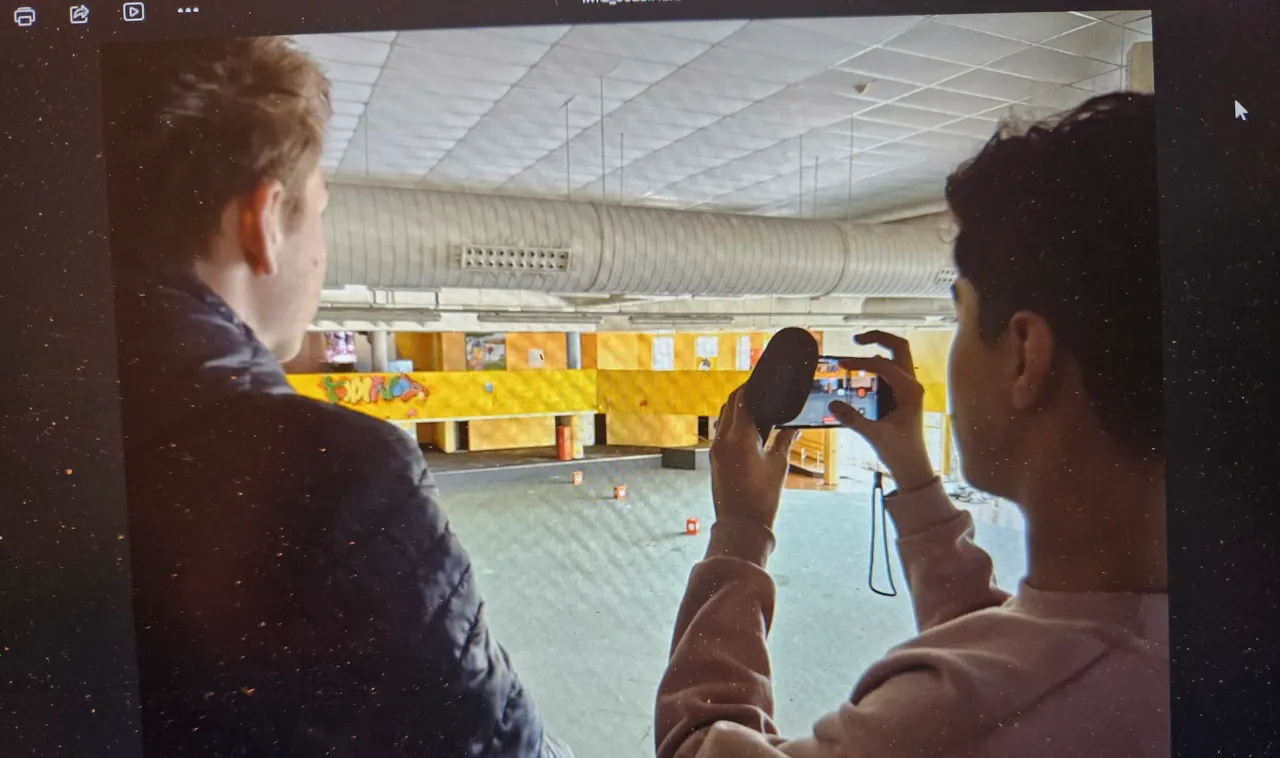 Zwei Jugendliche filmen in der großen, leeren Halle des ehemaligen Wellenbads. Im Hintergrund ist die gelb-grüne Wandgestaltung des früheren Indoor-Spielplatzes zu sehen.