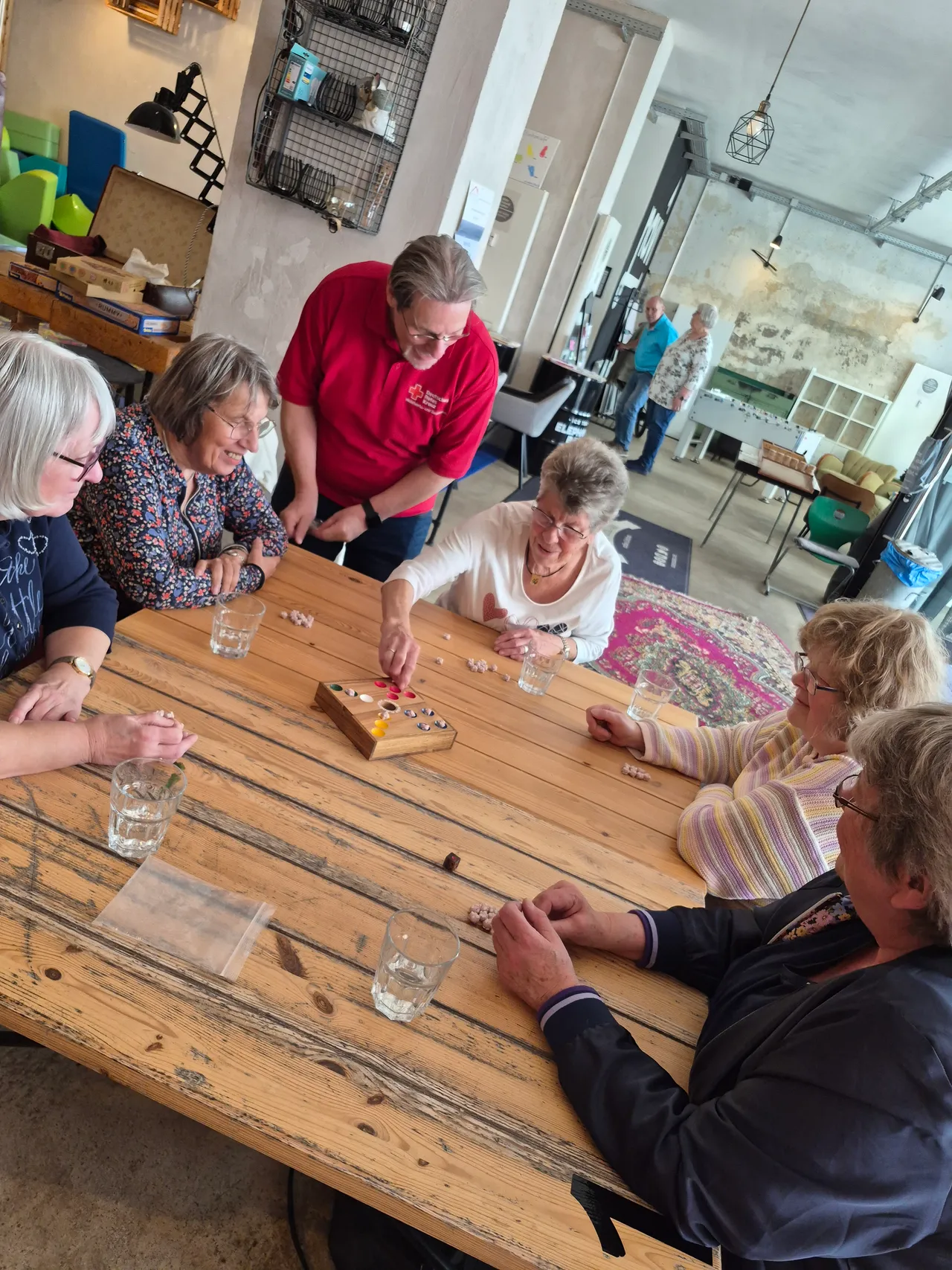 Mehrere ältere Frauen sitzen an einem Holztisch und spielen ein Brettspiel mit bunten Spielfiguren. Im Hintergrund sind weitere Personen und Möbel in einem hellen Raum zu sehen.