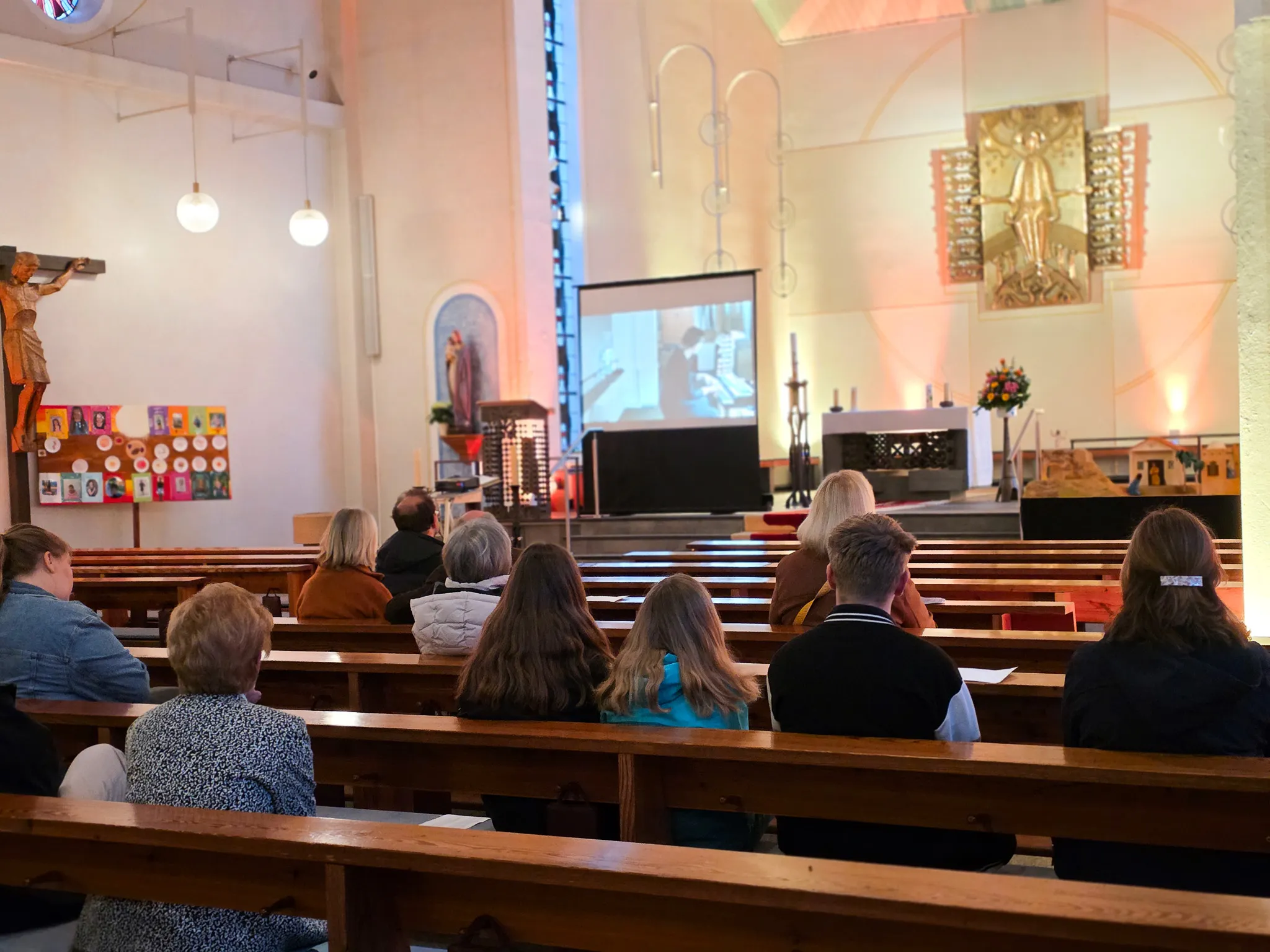 Zuschauer sitzen auf Kirchenbänken und schauen auf eine Großbildleinwand, auf der ein Organist zu sehen ist. Im Hintergrund ein Altar und religiöse Kunstwerke.