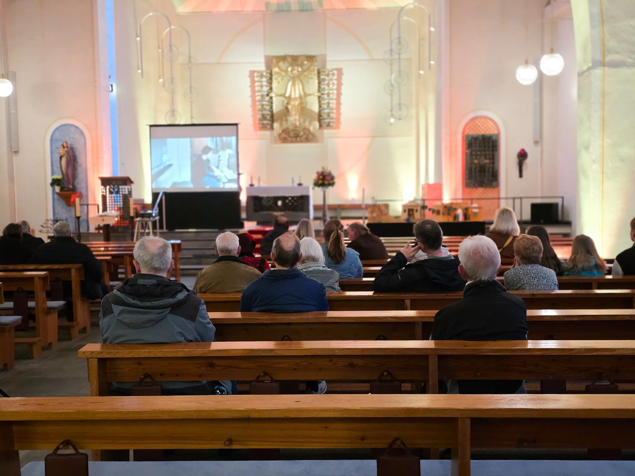 Verschiedene Menschen sitzen auf Kirchenbänken und schauen auf eine Großbildleinwand, auf der ein Organ zu sehen ist.