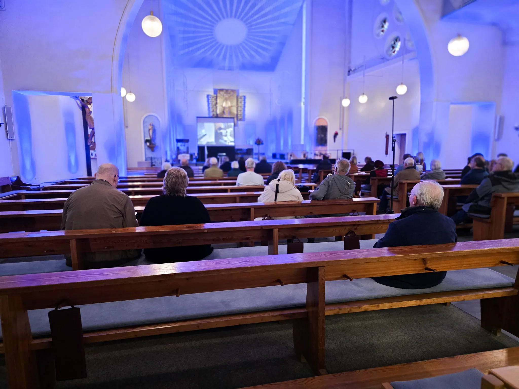 Verschiedene Menschen sitzen auf Holzbänken in einer Kirche und schauen in Richtung Altarbereich, wo eine Organistin spielt.