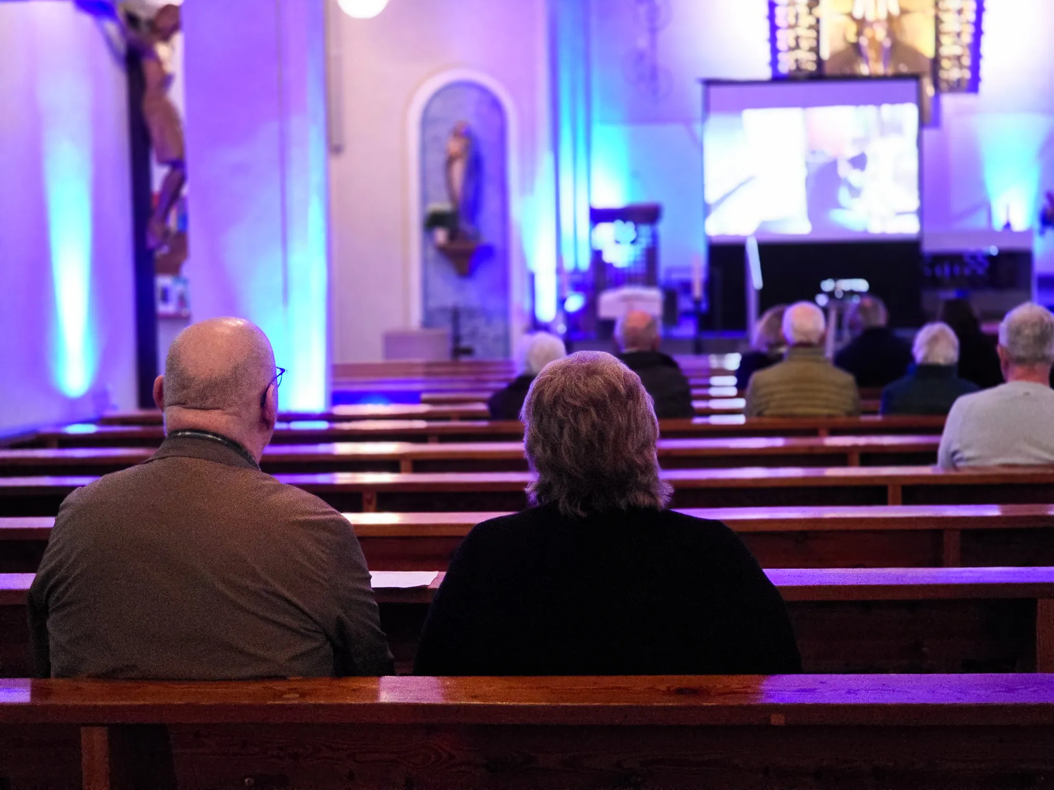 Zuschauer sitzen auf Holzbänken in einer Kirche und schauen in Richtung Altar und einer großen Leinwand, auf der ein Bild zu sehen ist. Im Vordergrund sind die Köpfe zweier Personen zu erkennen.