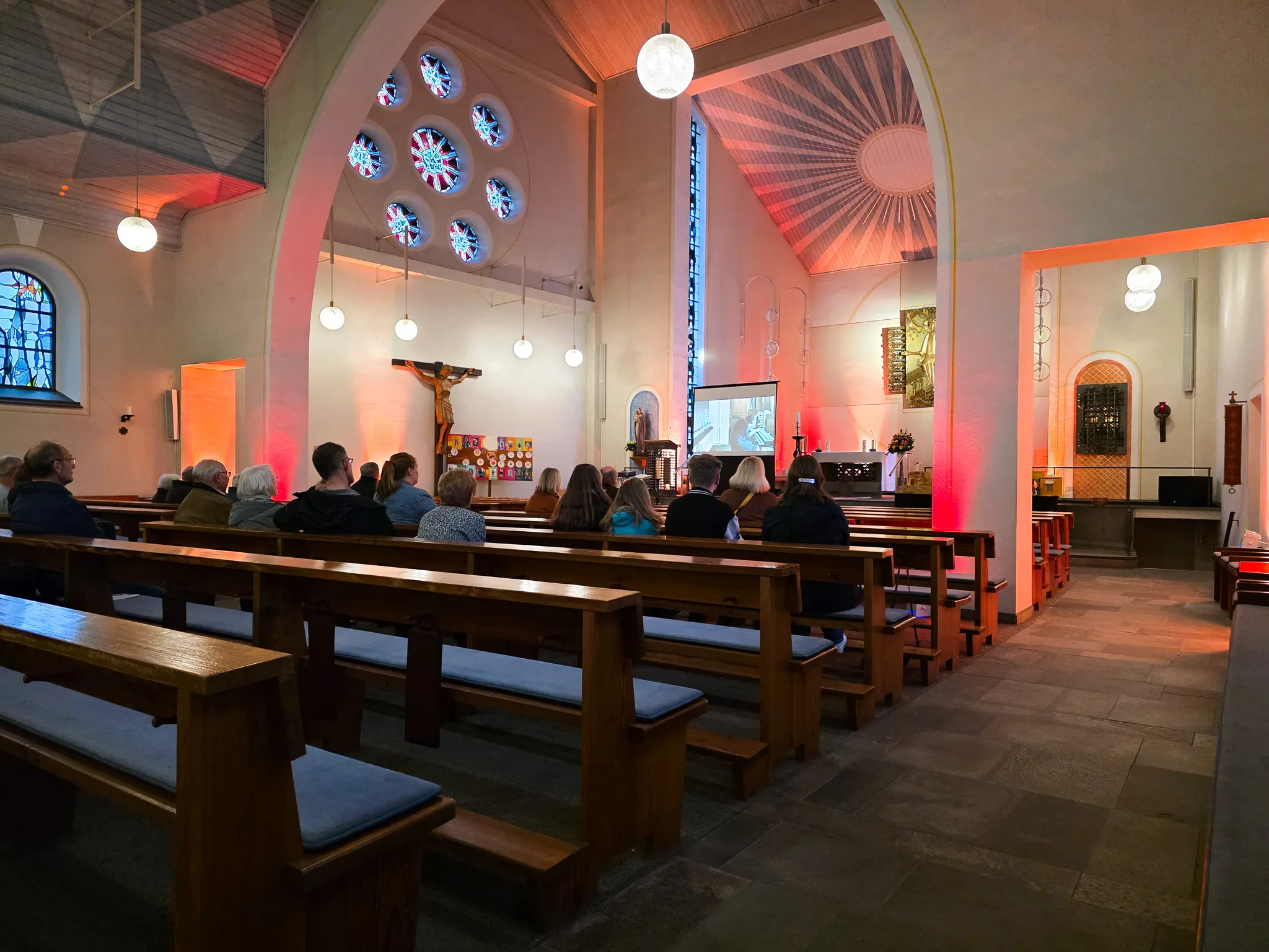Verschiedene Personen sitzen auf Holzbänken in einer Kirche und schauen auf eine Leinwand, auf der ein Bild zu sehen ist. Im Hintergrund ein Kruzifix und farbige Glasfenster.
