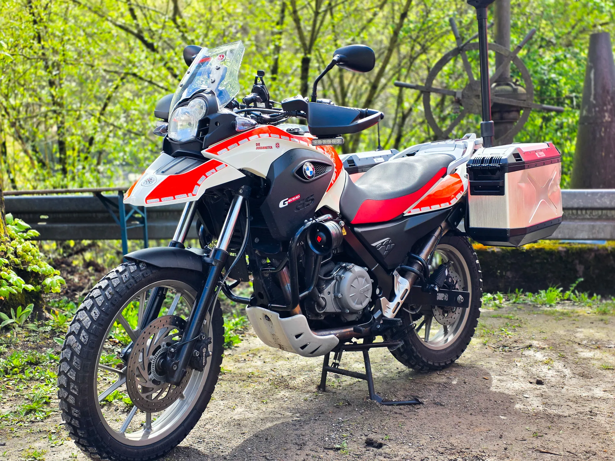 Rote und weiße BMW-Geländemotorrad der Polizei mit Gepäckträger steht auf einem Parkplatz vor Büschen und einer Holzbank.