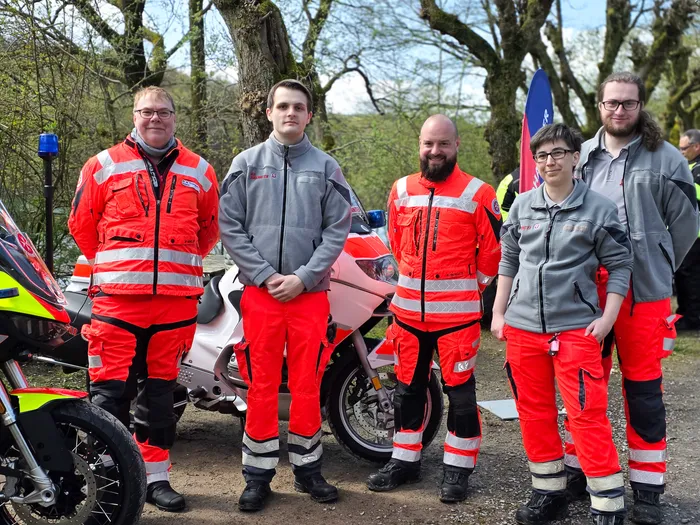 Fünf Personen in Warnwesten und roter Motorradbekleidung stehen vor Motorrädern, im Hintergrund eine Flagge und Bäume.
