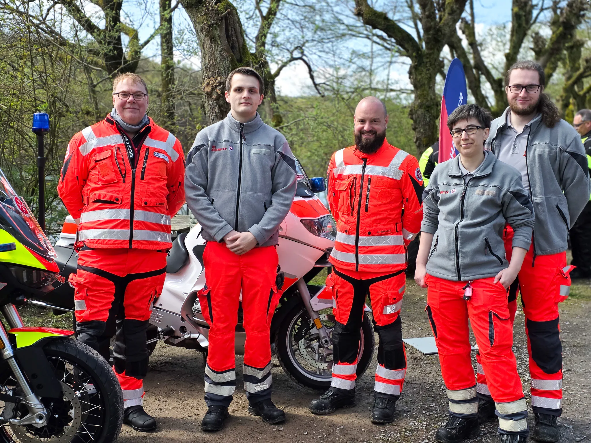 Fünf Personen in Warnwesten und roter Motorradbekleidung stehen vor Motorrädern, im Hintergrund eine Flagge und Bäume.