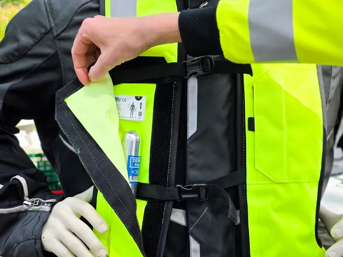 Eine Hand öffnet eine Tasche an einer gelben Warnweste, in der eine Erste-Hilfe-Ausrüstung und eine kleine Gaspatrone zu sehen sind.