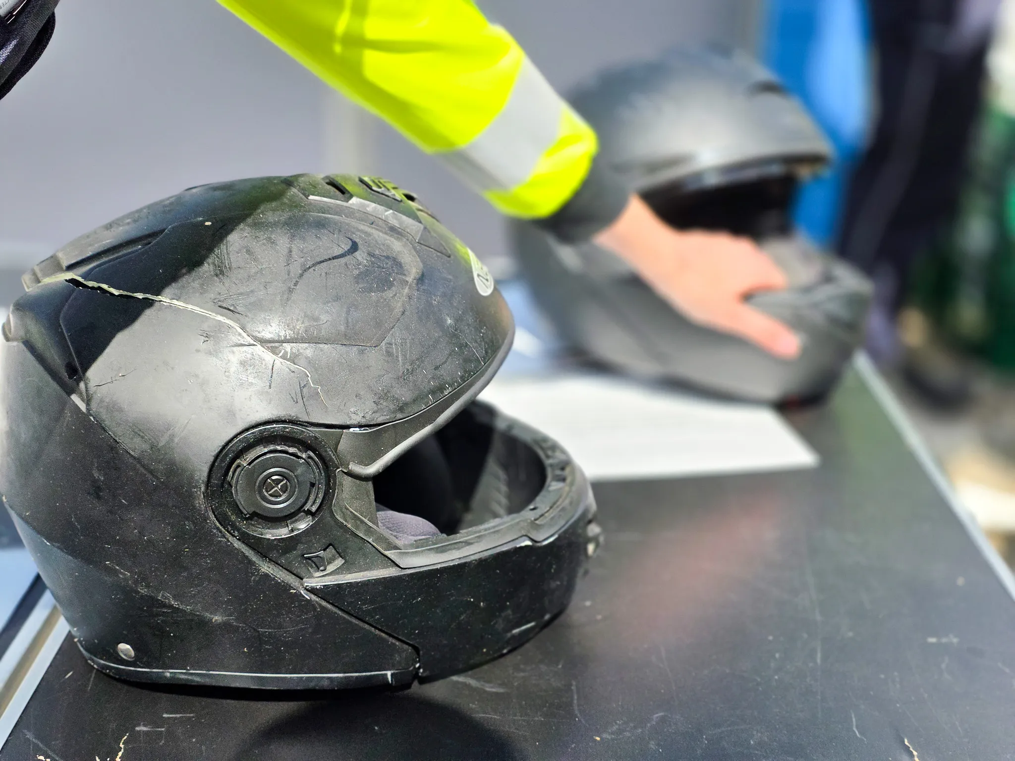 Beschädigter Motorradhelm liegt neben weiteren Helmen auf einer dunklen Oberfläche, im Hintergrund ein Polizist mit Warnweste.
