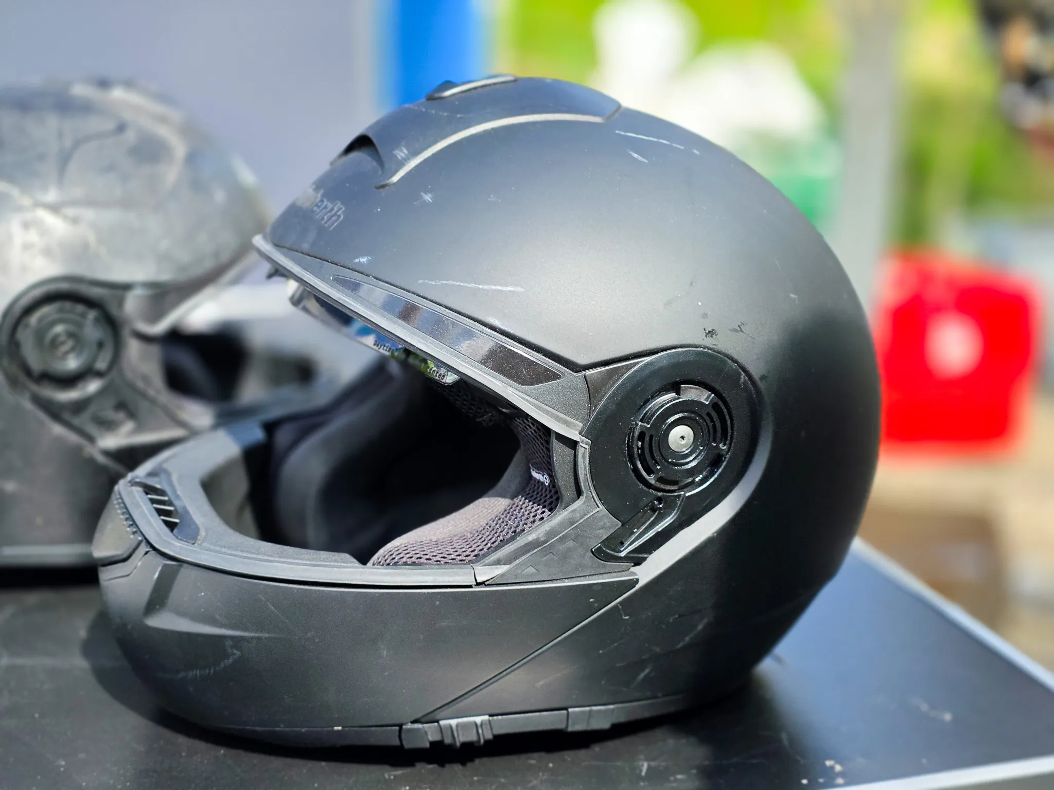 Schwarzer Motorradhelm mit aufklappbarem Visier auf einer dunklen Oberfläche, im Hintergrund verschwommene Farben und Formen.