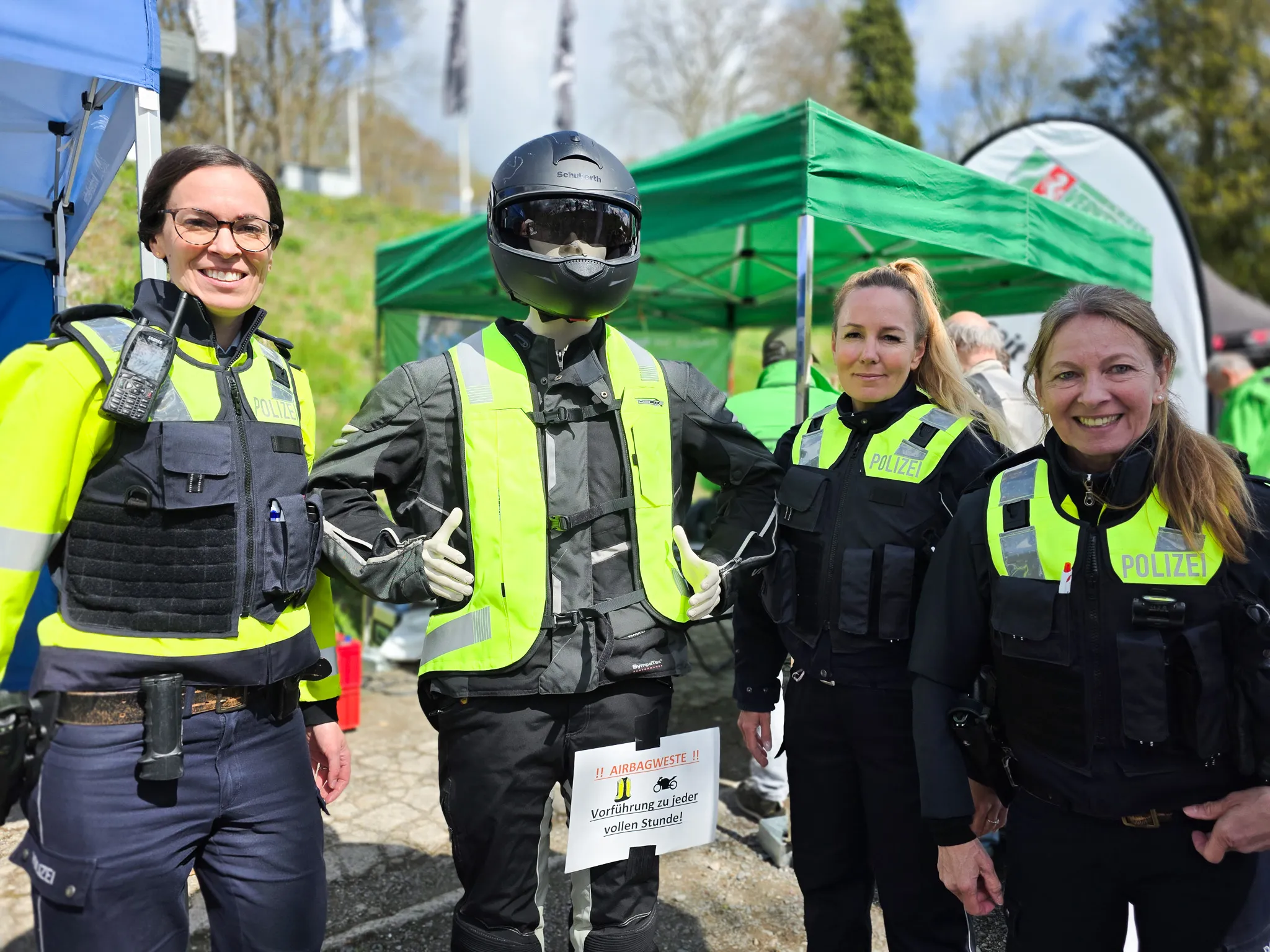 Drei Polizistinnen und ein Dummy in Motorradmontur stehen vor Zelten und Bäumen. Das Schild vor dem Dummy weist auf Airbagwesten und regelmäßige Vorführungen hin.