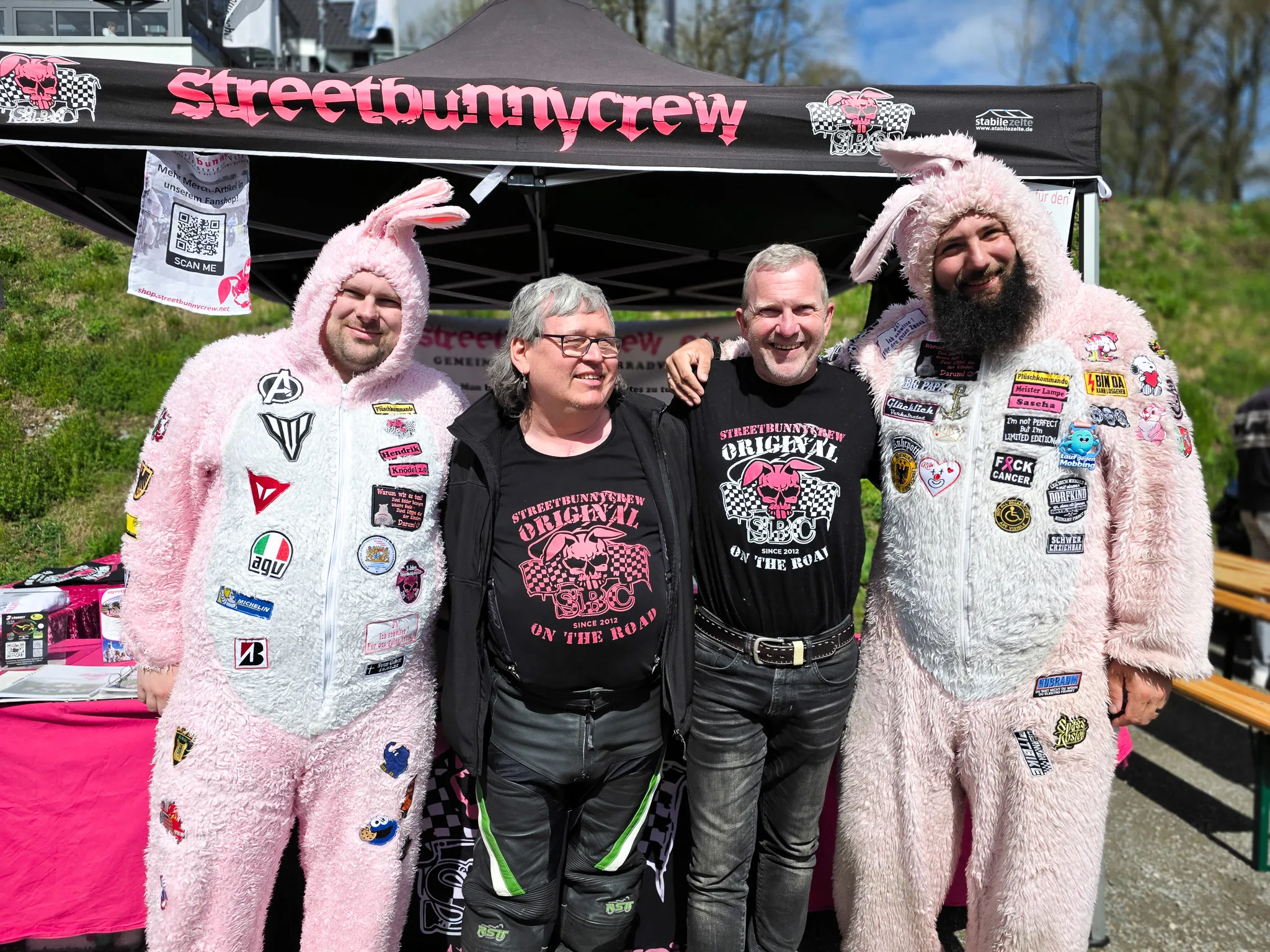 Vier Personen stehen vor einem Verkaufsstand mit dem Aufdruck „Streetbunnycrew“. Die beiden äußeren Personen tragen rosa Hasenkostüme, die mittlere Person trägt ein schwarzes Shirt mit einem Motorrad-Design und die vierte Person ein schwarzes Shirt mit Totenkopf-Motiv.